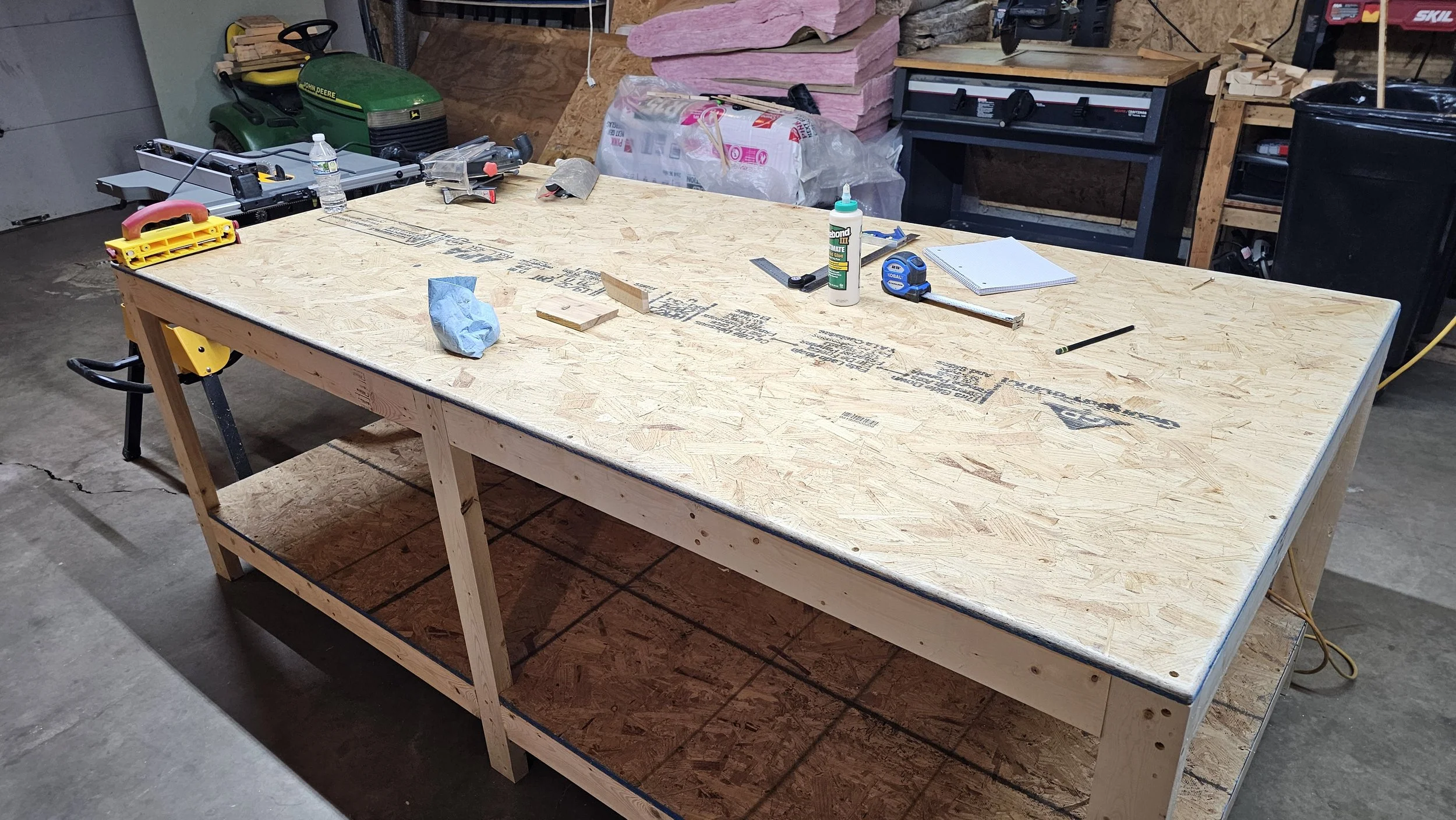 A woodworking workbench in a garage, with various tools and supplies on top, including a water bottle, a glue bottle, a tape measure, a notepad, and small pieces of wood. In the background, there is a John Deere lawn tractor, pink insulation, and sto
