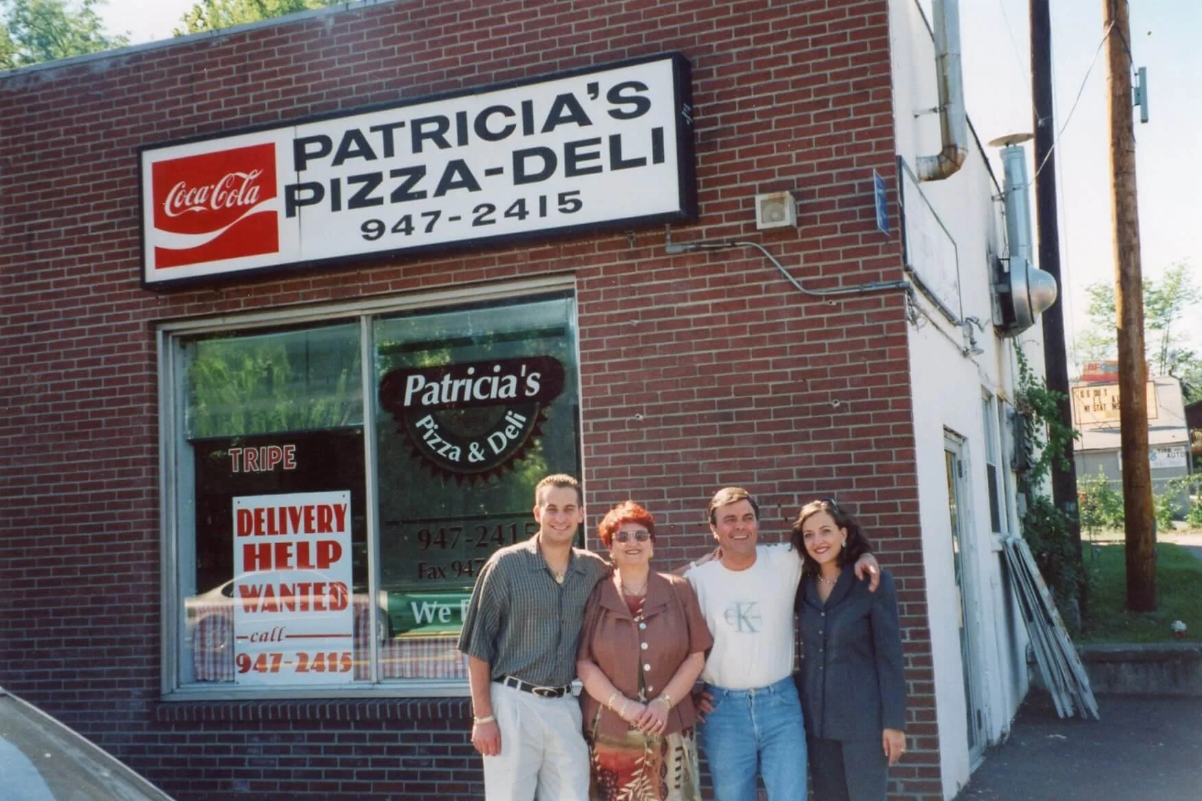 Chef Joey and Chef Tomaso standing with their family in front of a brick pizza restaurant named Patricia's Pizza & Deli.