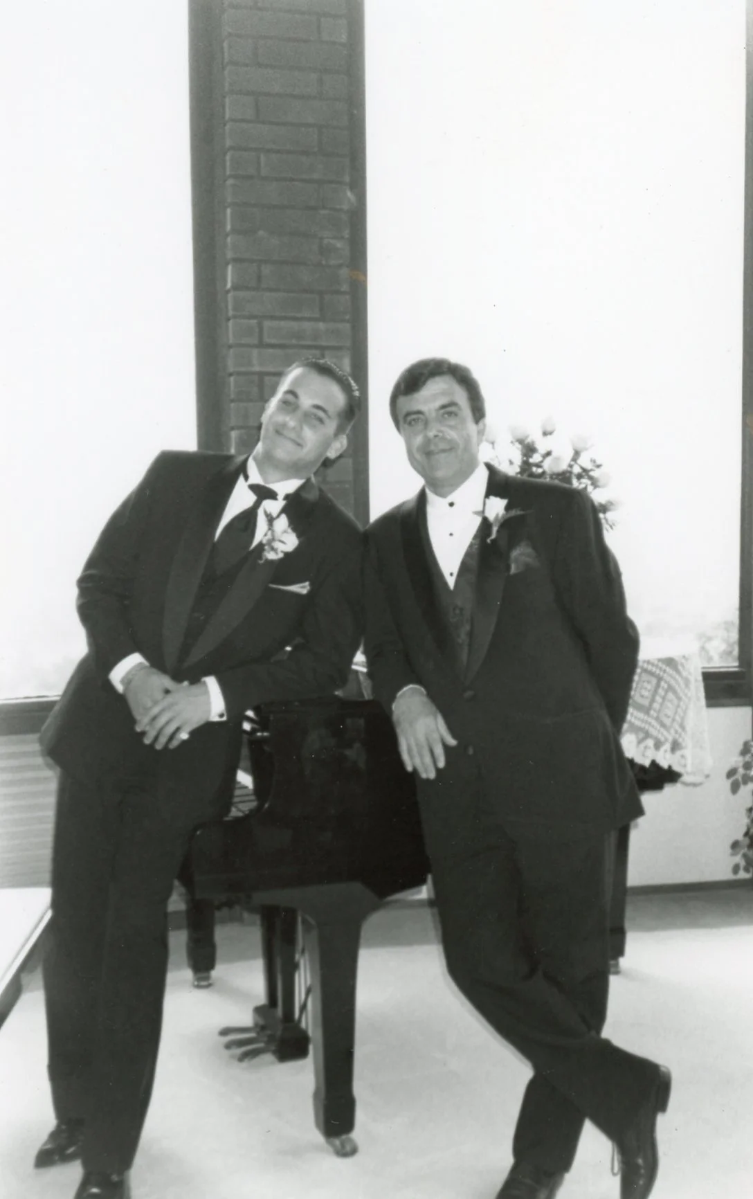 Chef Joey and Chef Tomaso in tuxedos standing next to a piano in a room with large windows, brick wall, and flowers in the background.