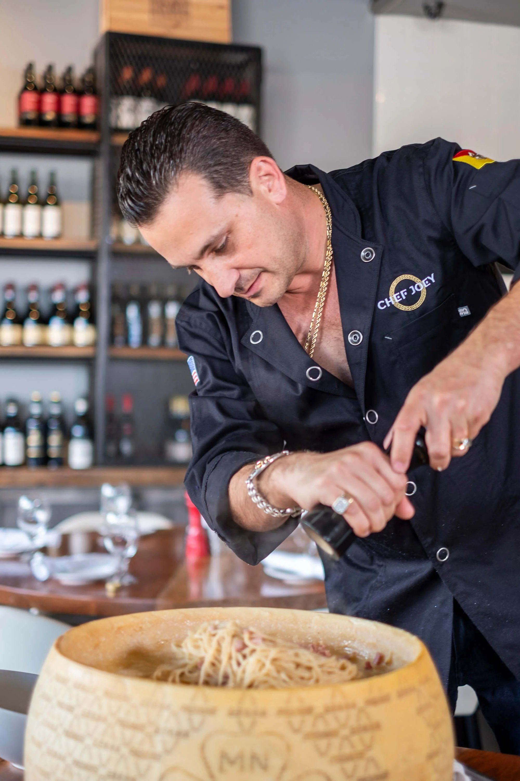 A man with dark, slicked-back hair and wearing a black chef's coat, gold chains, and rings is preparing food in a restaurant. In the foreground, there is a large wooden bowl filled with pasta. Shelves with bottles and jars are visible in the background.