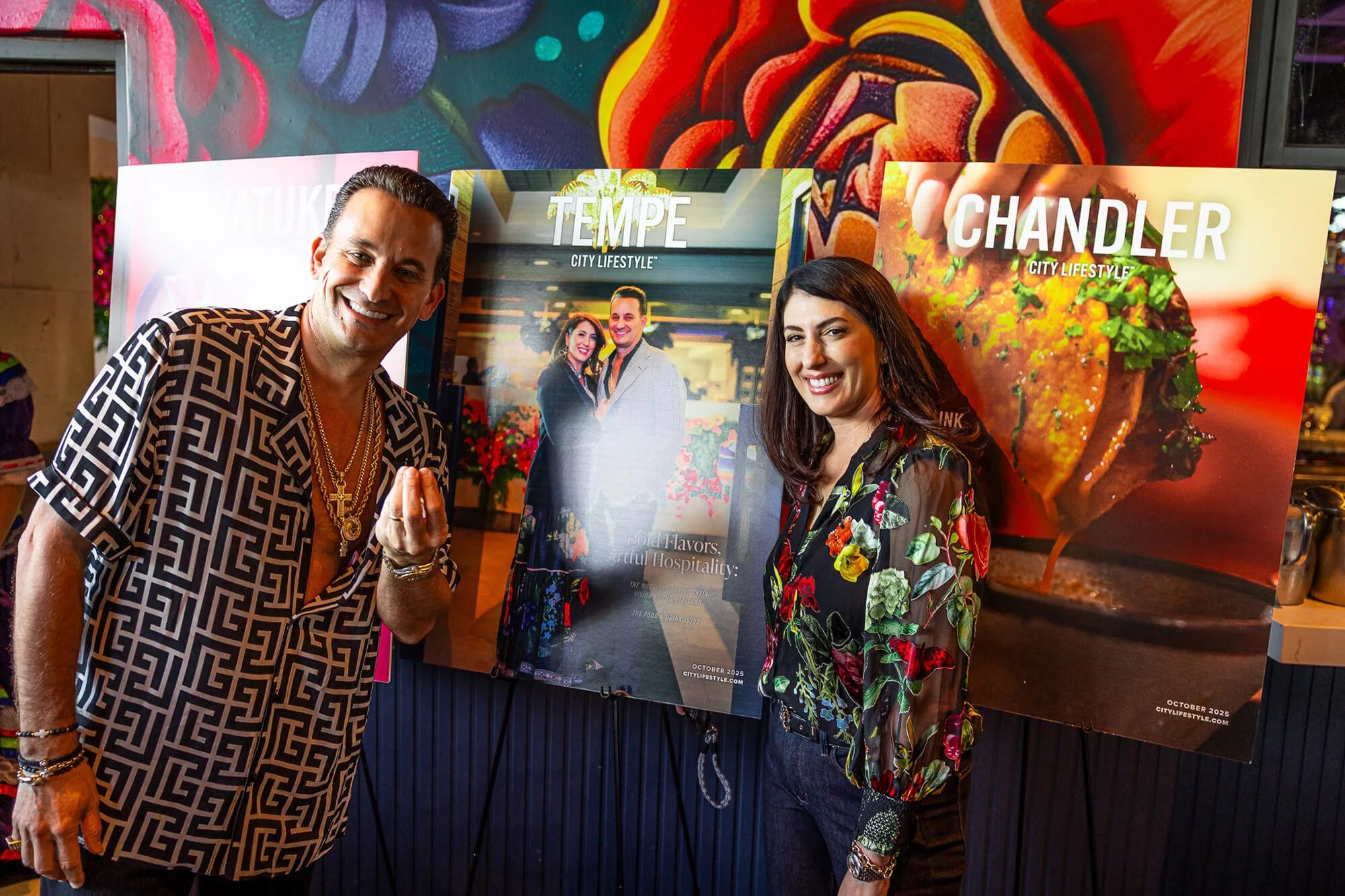Two people smiling at the camera, standing in front of restaurant concept opening posters that read 'TEMPE' and 'CHANDLER.' The man on the left is Chef Joey Maggiore, with his wife on the right Cristina Maggiore