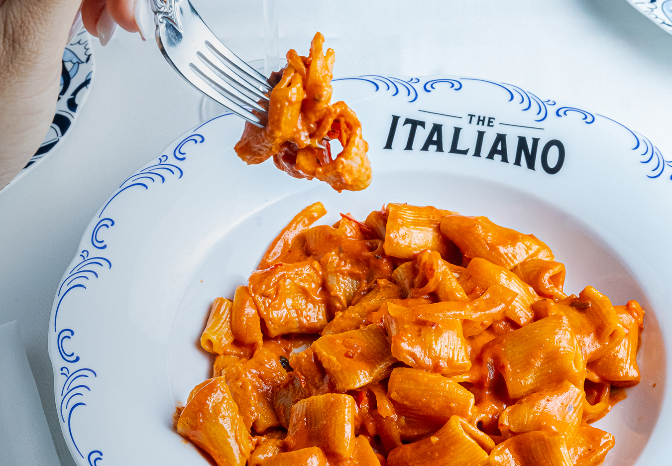The Italiano: A white plate with blue decorative borders and the word 'The Italiano' printed on it, containing pasta with red-orange tomato sauce. A hand is holding a fork with a bite of pasta.