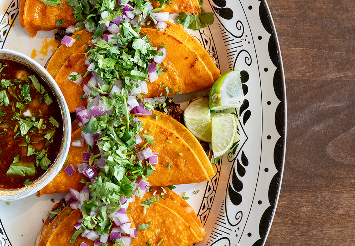 The Mexicano: Close-up of three beef tacos with chopped onions and cilantro on a decorative black and white plate, with lime wedges and a bowl of salsa on the side.