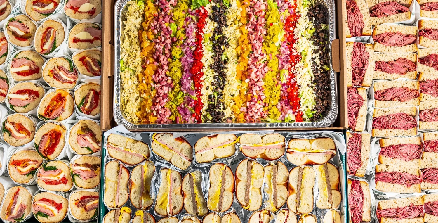 The Delicatessen: Assorted sandwiches, including mini wraps, and a colorful layered chopped salad in a large tray.