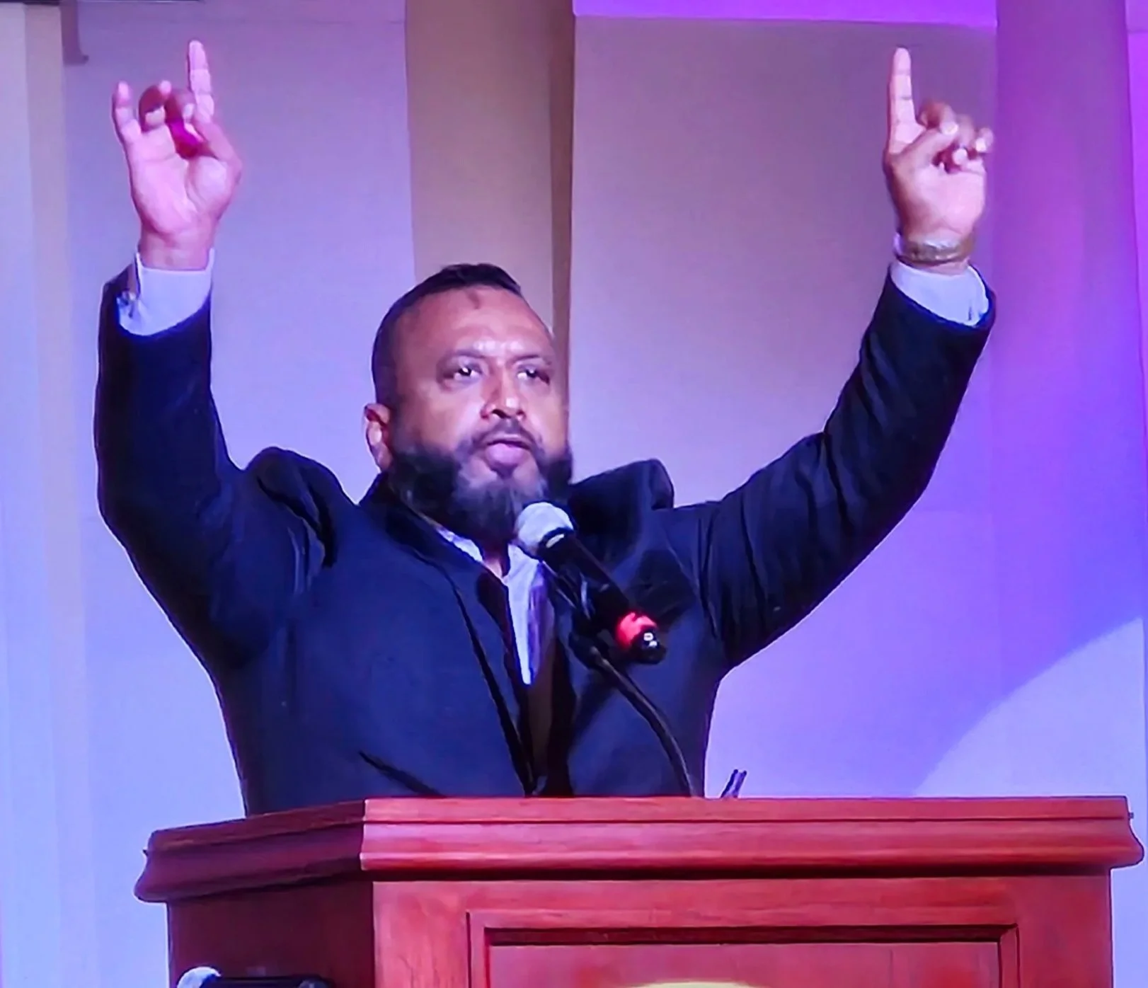 A man in a suit is standing at a podium with his right arm raised, pointing towards the ceiling, during a formal event or speech. There are purple and pink lighting effects behind him.