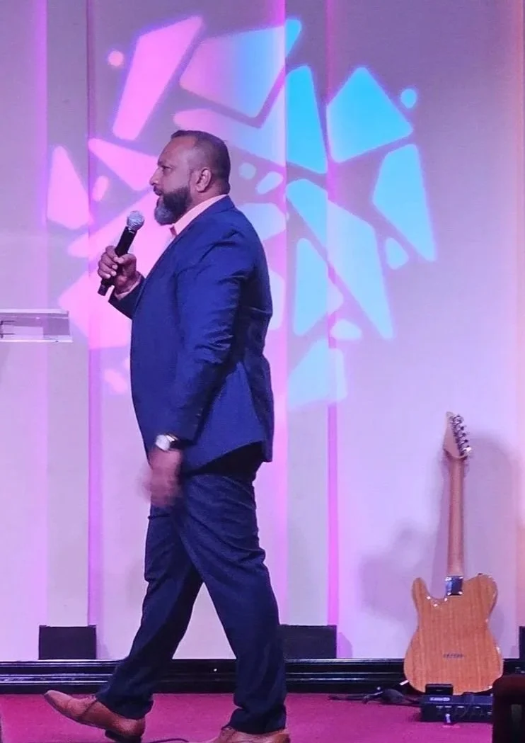 A man in a dark blue suit and pink shirt speaking into a microphone on stage at an event with the letters 'EAG' on the podium, in Houston, TX, with a diverse audience seated facing him. The stage has purple and blue decorative lighting, a small clear