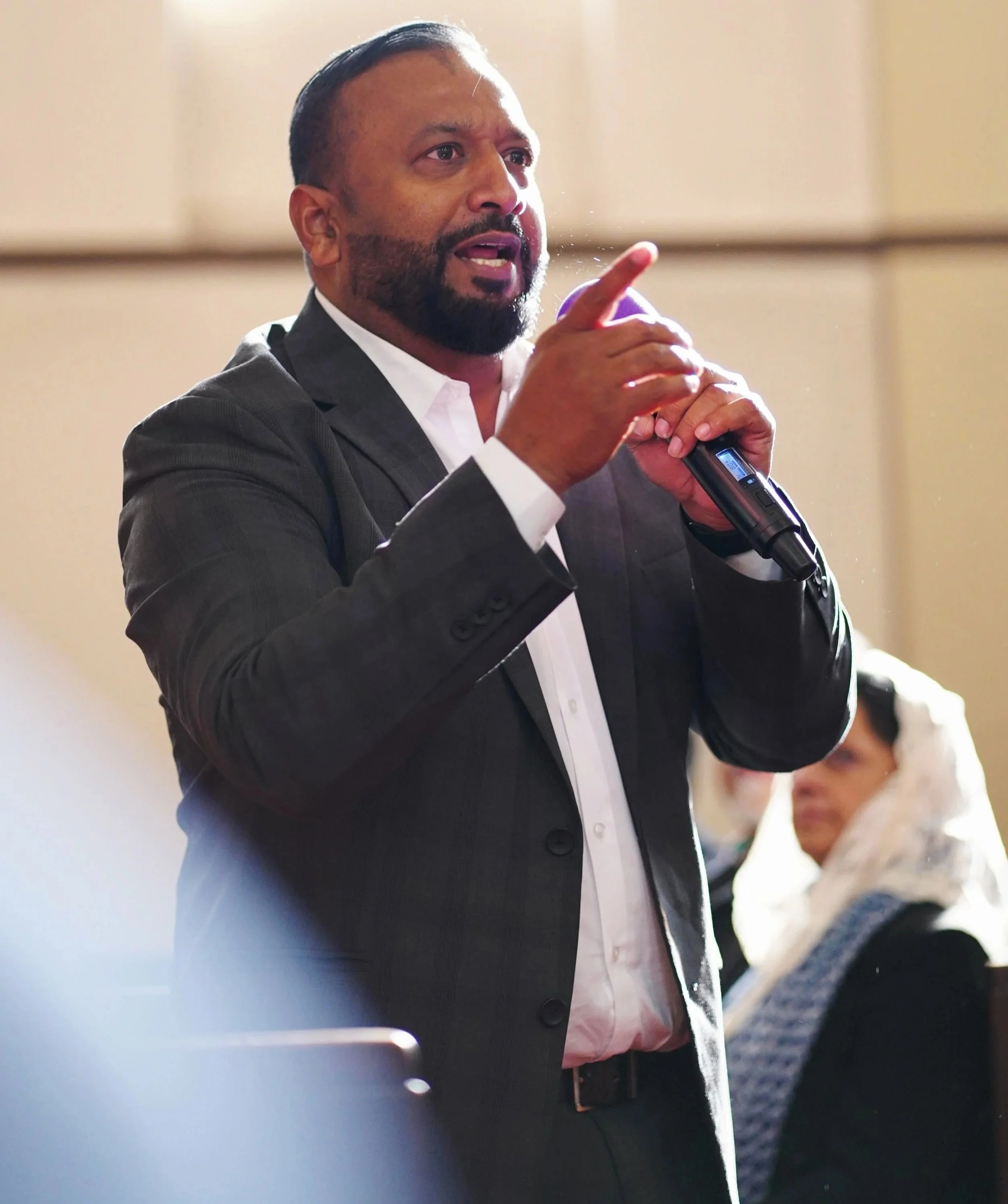 A man in a suit is speaking, holding a microphone, and gesturing with his hand. There are people in the background, including a woman listening.