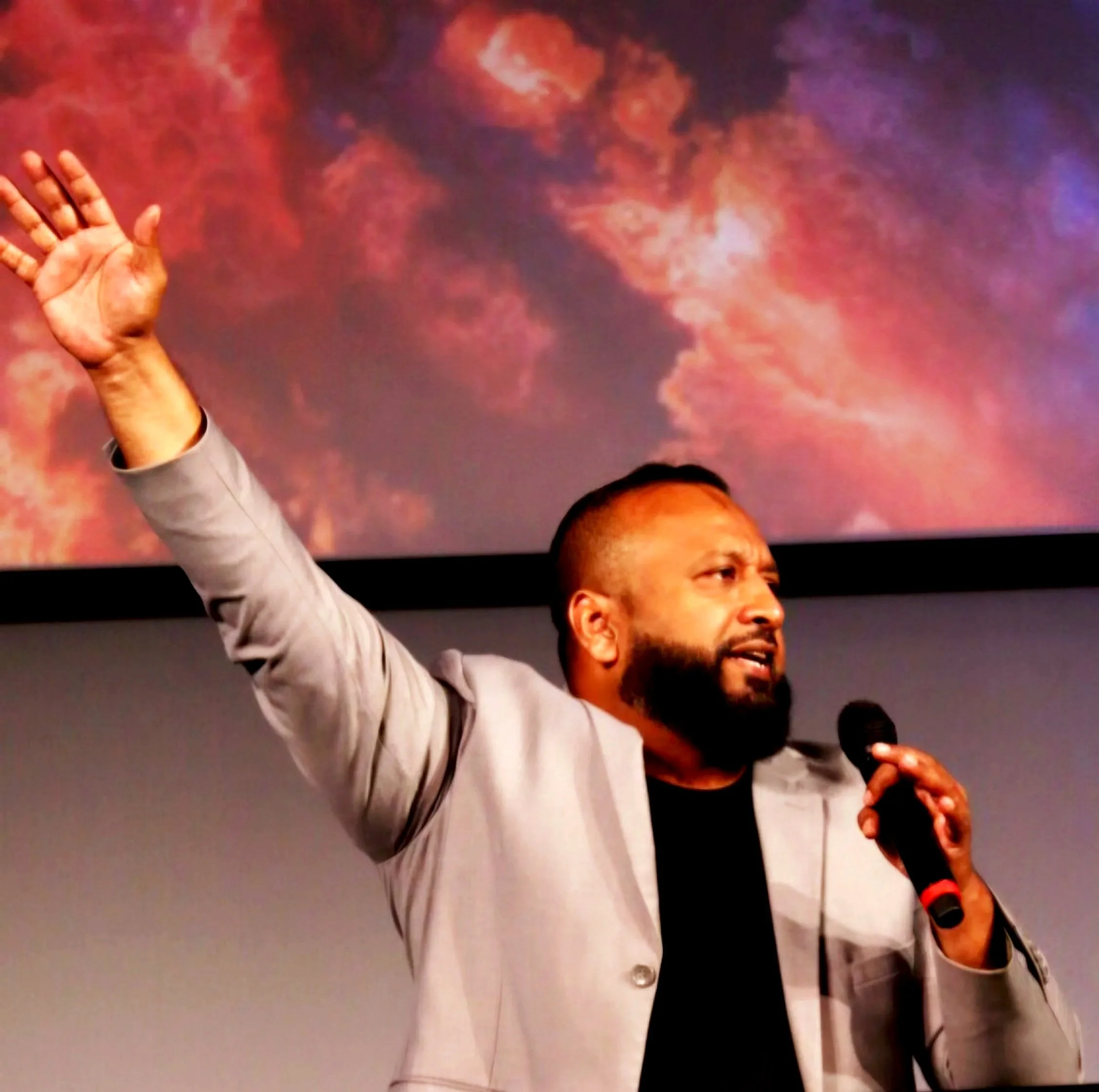 A man in a gray suit and white shirt is raising his hand, possibly speaking or greeting, with a colorful, cloud-like background. Part of another person's head is visible at the bottom of the image, and the man holds a microphone in his other hand.