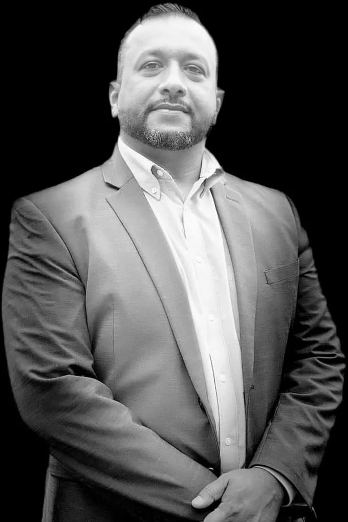 A man in a suit with a beard and short hair, standing with arms crossed, in front of a black background.