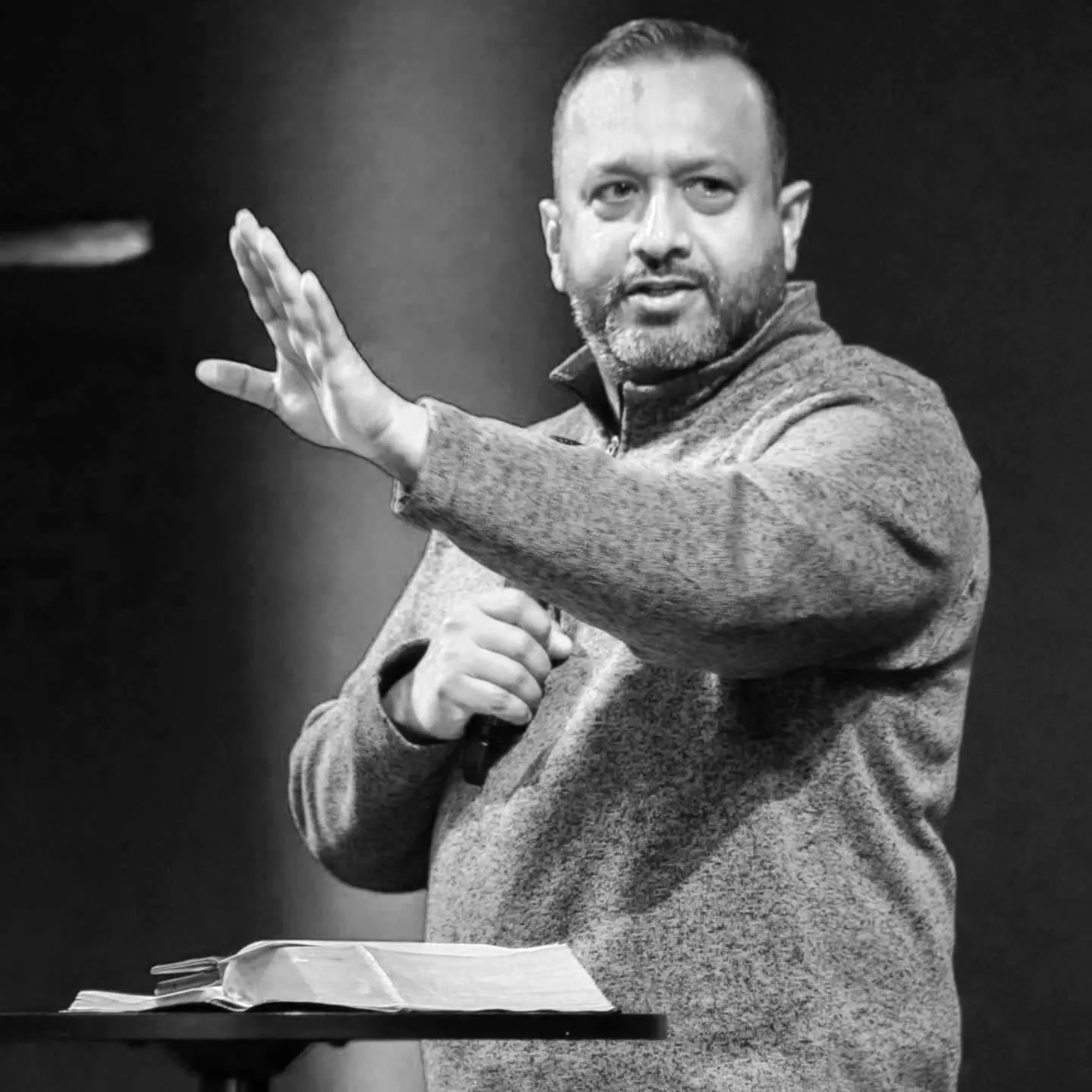 A man speaking or giving a presentation in front of a microphone, with an open book or notebook on a table in front of him.