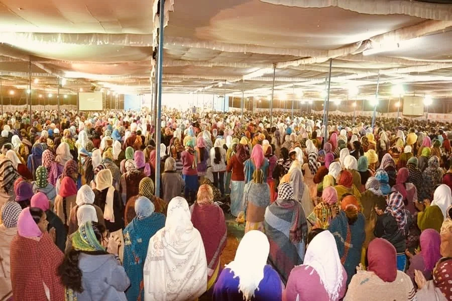 A large indoor gathering with many people, mostly women, wearing colorful headscarves, under a tent-like structure with hanging lights and projection screens.