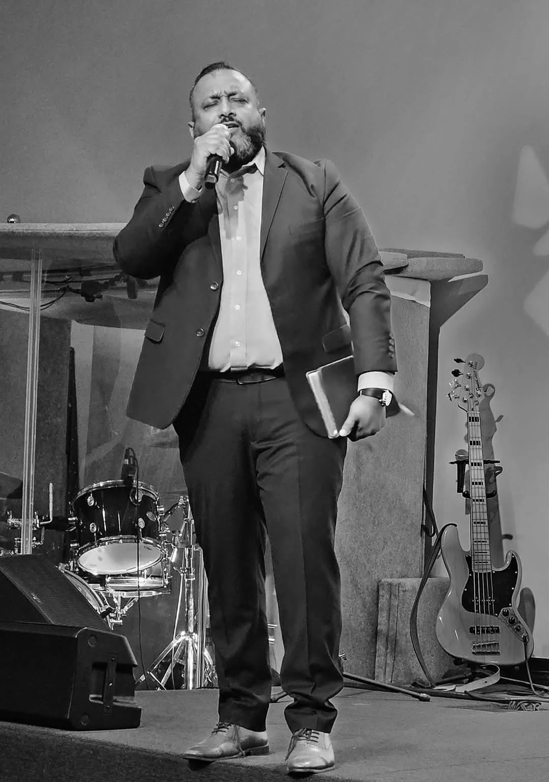 A man in a suit singing into a microphone on stage, holding a notebook, with musical instruments including a bass guitar, drum set, and keyboard in the background.