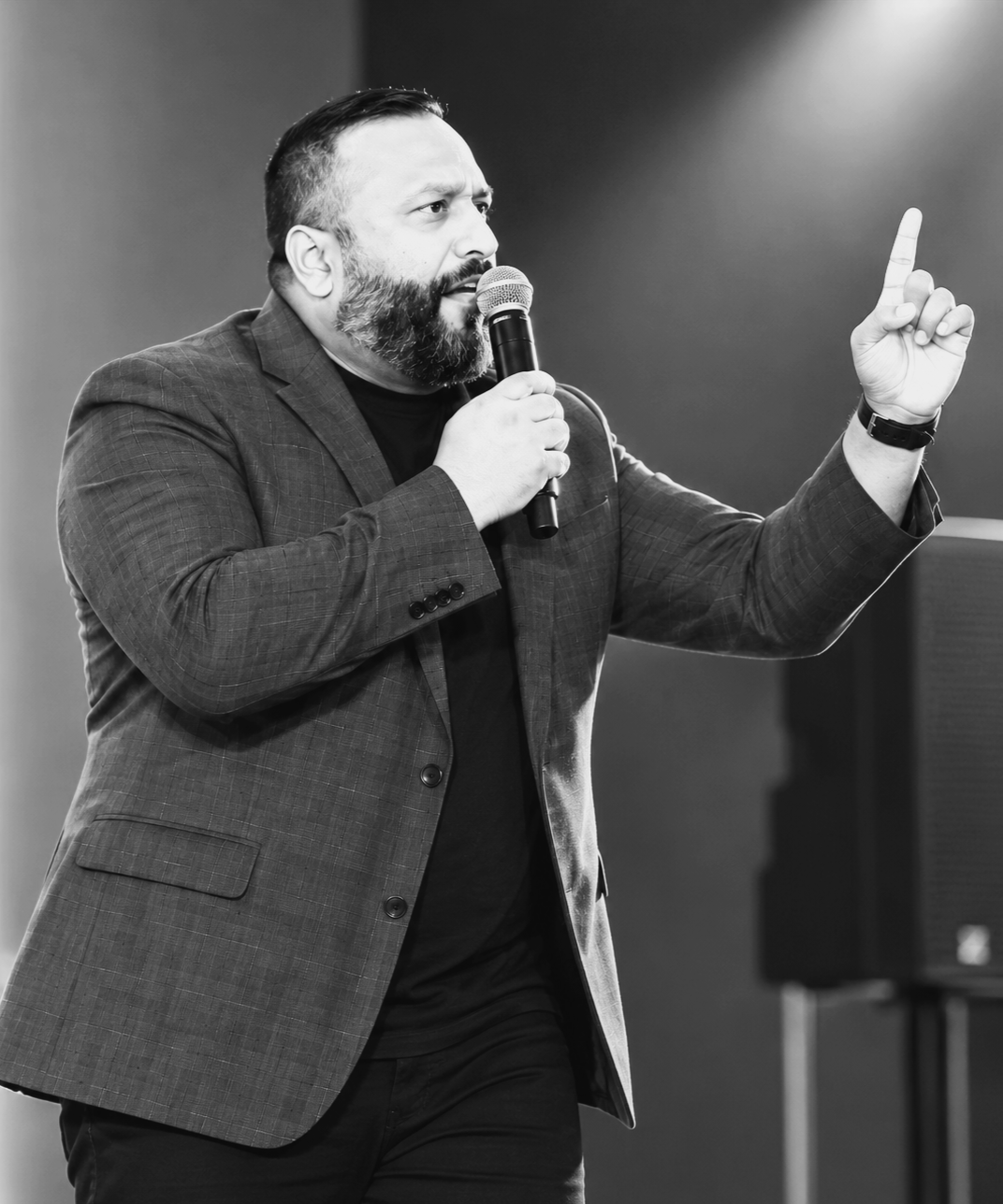 A man with a beard wearing a blazer and black shirt, holding a microphone and gesturing with his right hand, appears to be giving a speech or presentation indoors.