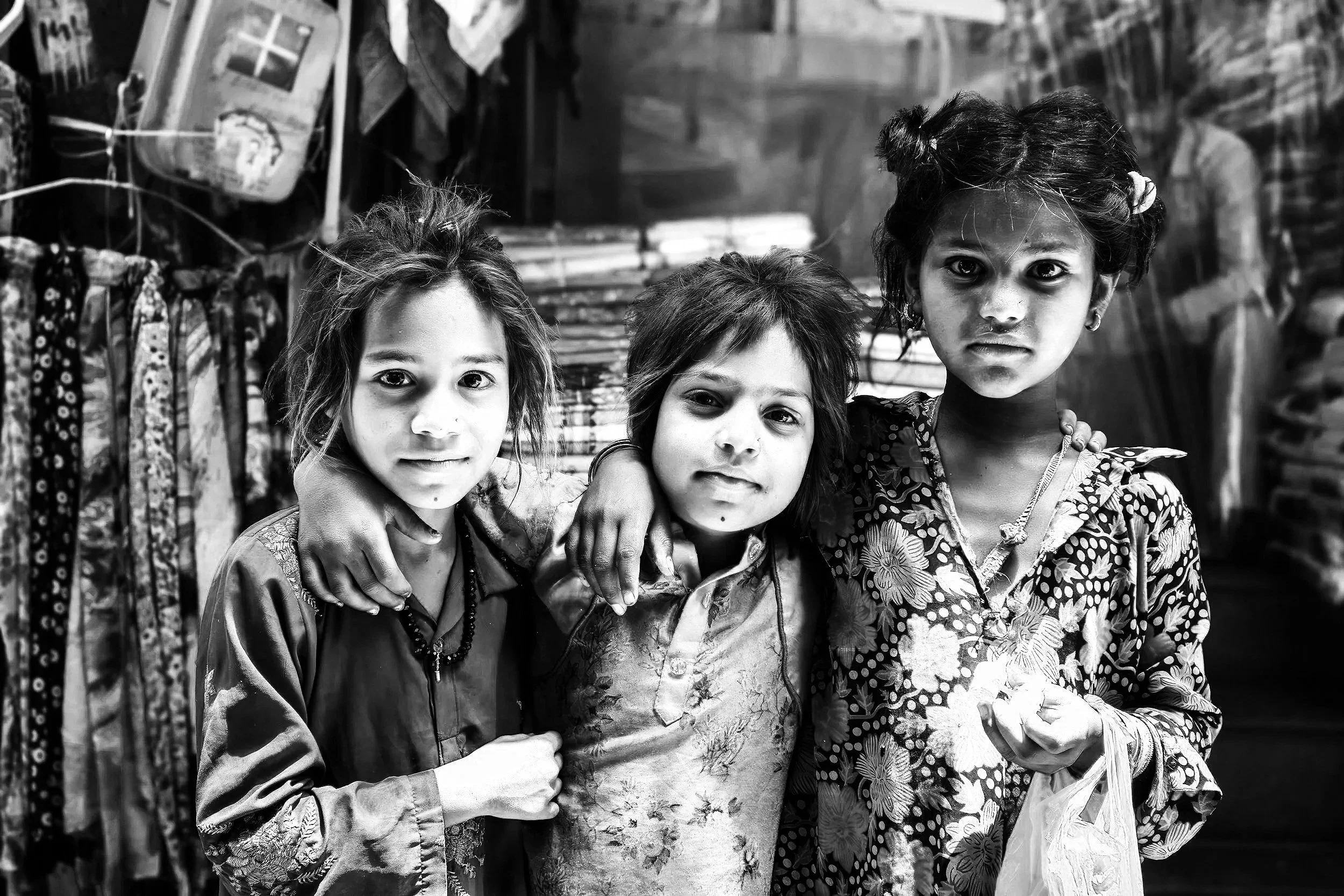 Three women standing closely together with their arms around each other in a marketplace, surrounded by hanging clothes and fabric.