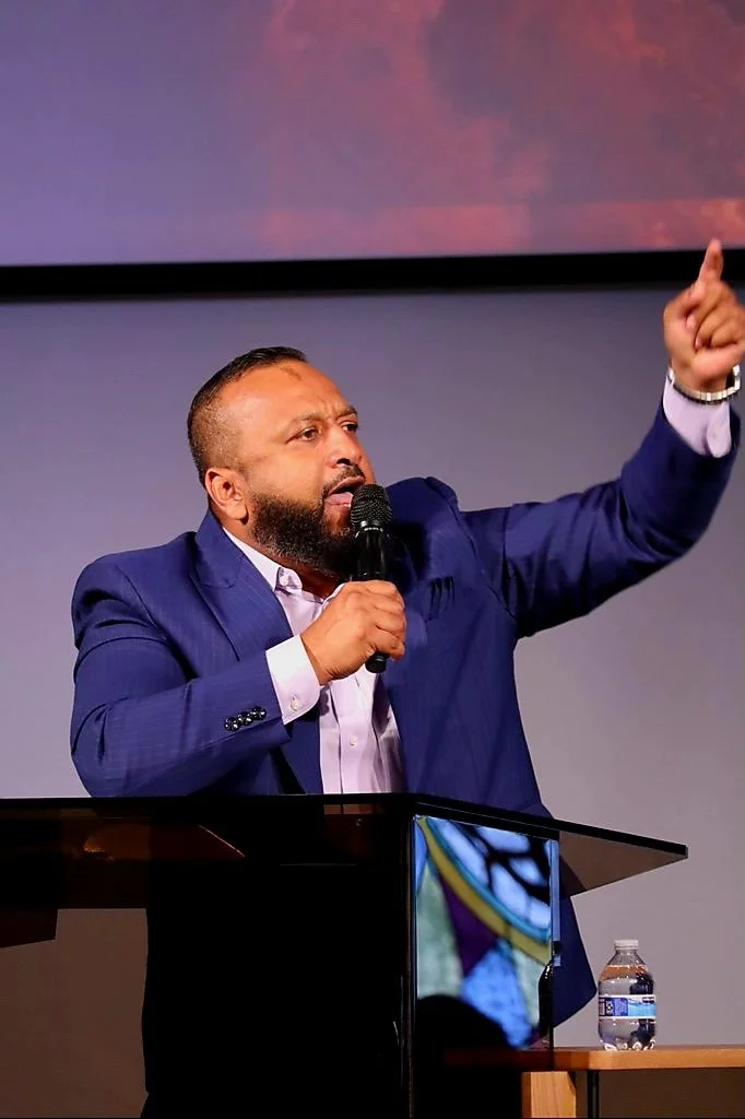 Man in a blue suit giving a speech at a podium, holding a microphone and gesturing with his right hand.