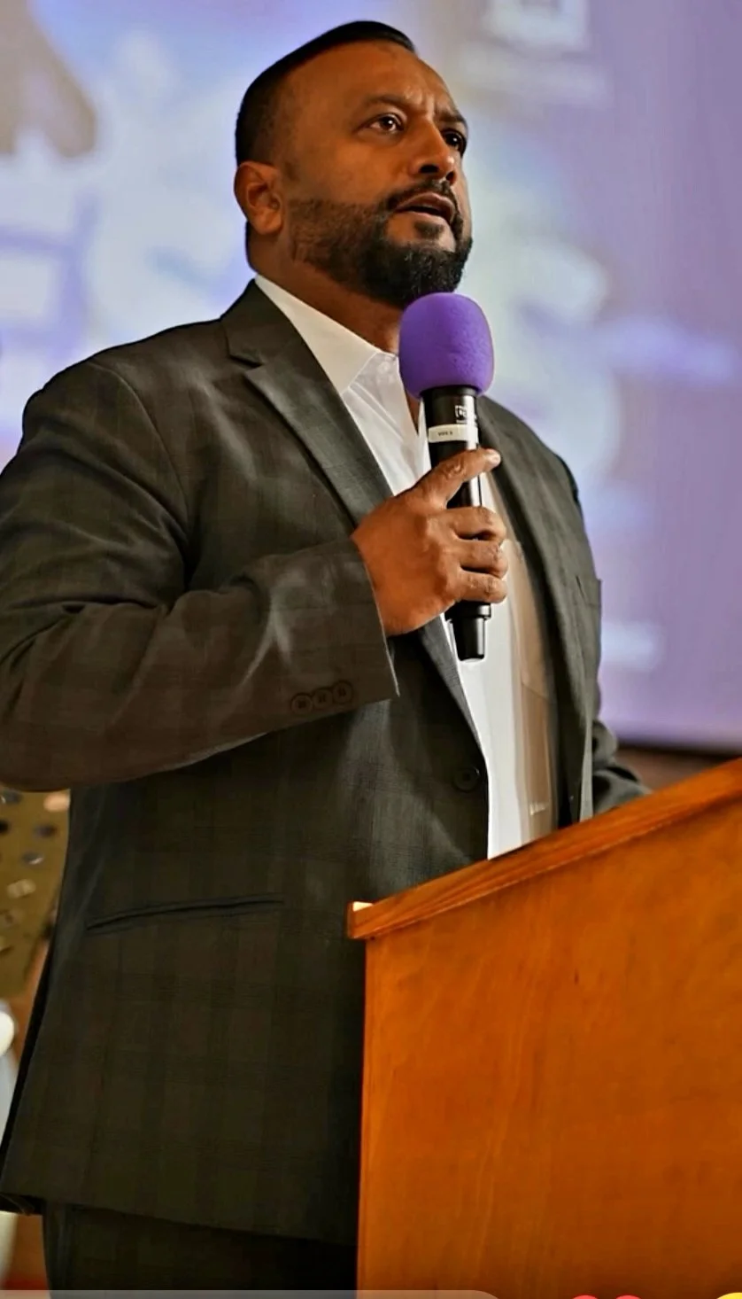 A man in a dark suit and white shirt standing behind a wooden podium, holding a purple microphone during a speech or presentation.