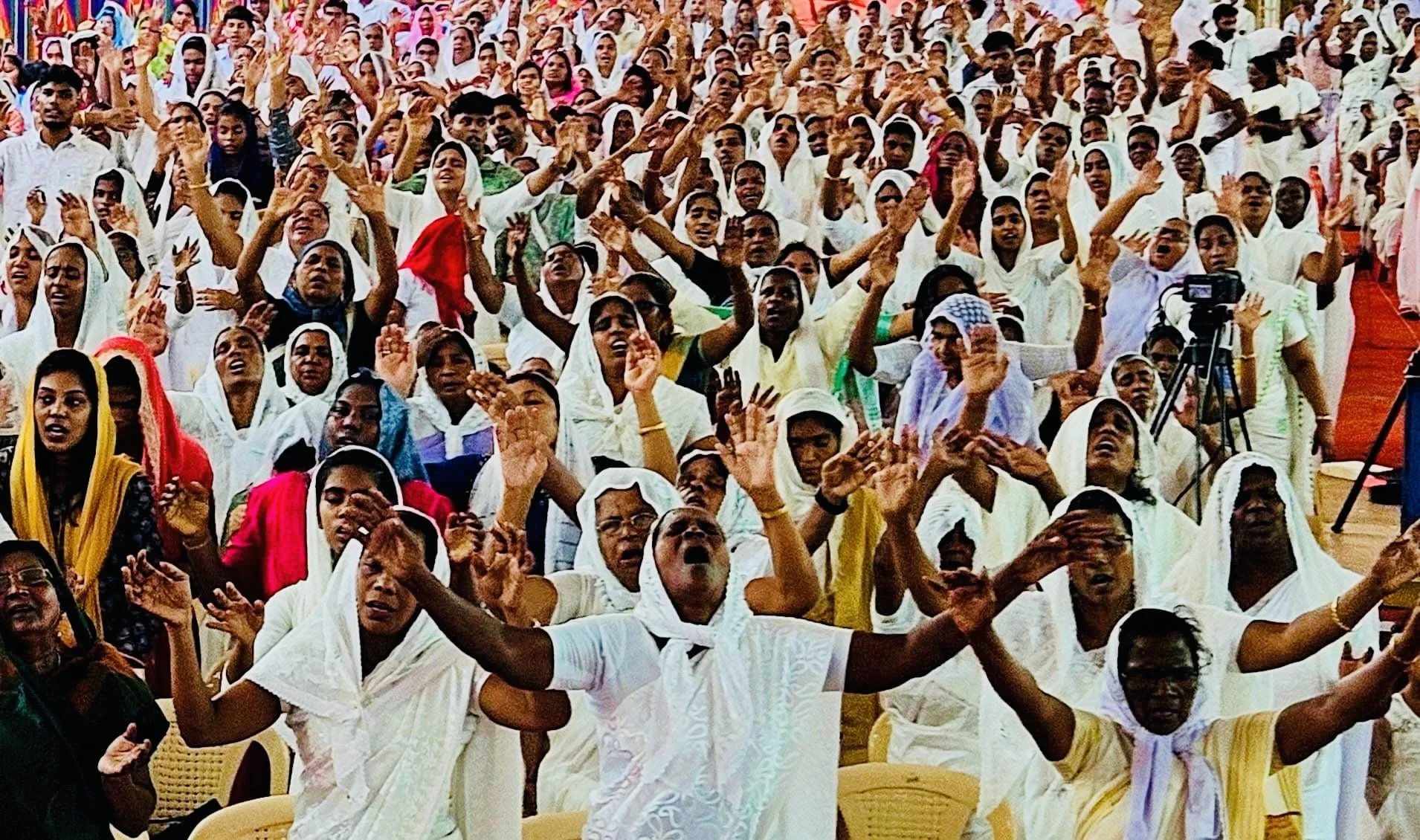 Large gathering of women dressed in white, some with head coverings, raising their hands and worshipping.