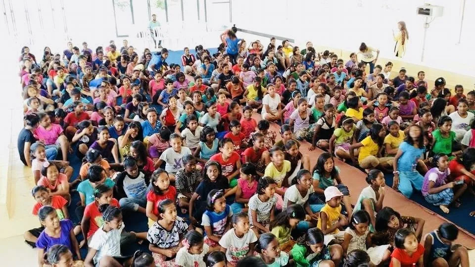 A large group of children seated on the floor in a bright room, some adults standing or sitting at the edges of the room.
