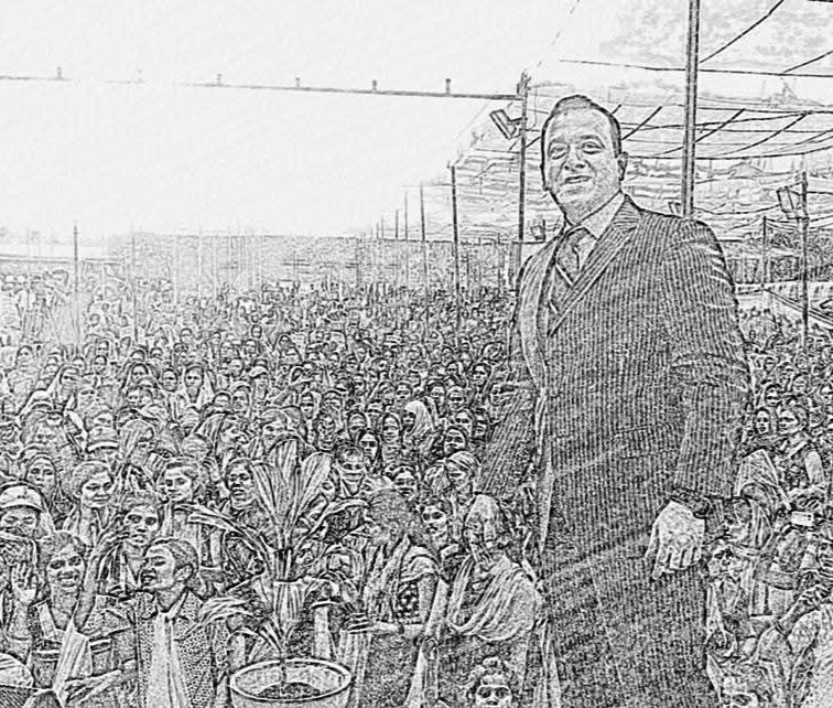 A man in a business suit standing among a large crowd of people, with some outdoors in an open area with structures overhead.