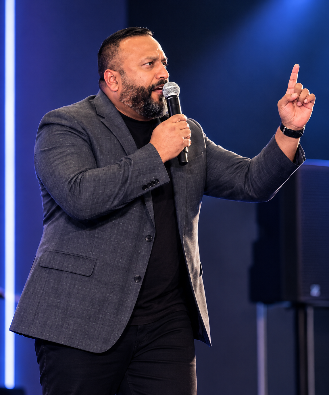 Man in a gray suit with a black shirt holding a microphone and gesturing with his right hand, speaking at an indoor event with blue lighting.