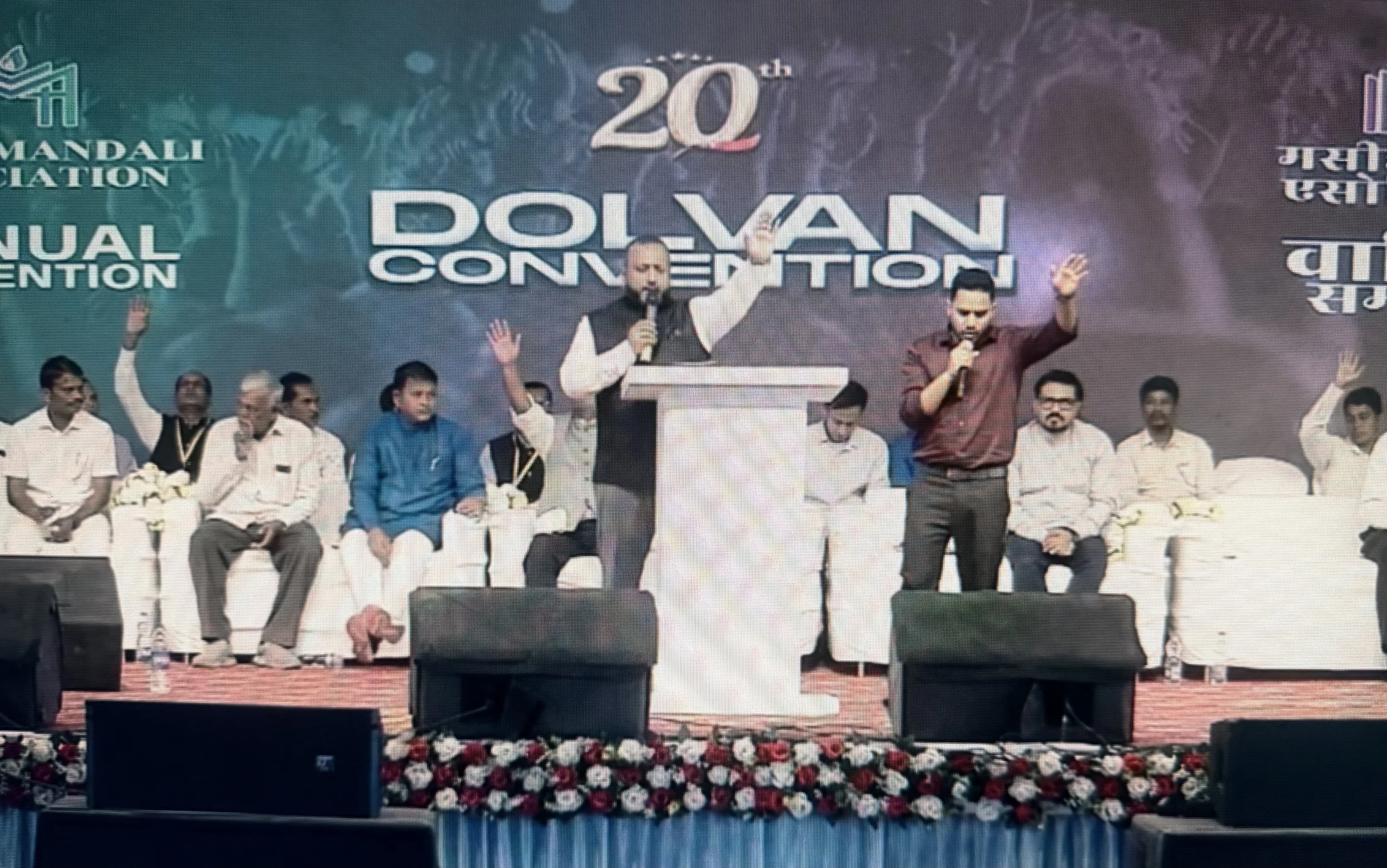 A group of people on stage at the 20th Dalvan Conference, with two men standing at the center speaking into microphones, and others seated behind them. The stage has a backdrop with text indicating the event and a floral decoration in front.