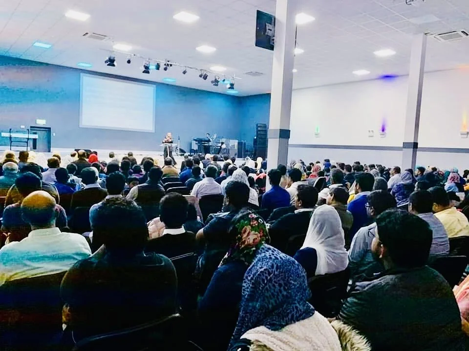 A large audience seated in a conference room, facing a stage with a speaker at a podium and a presentation screen.