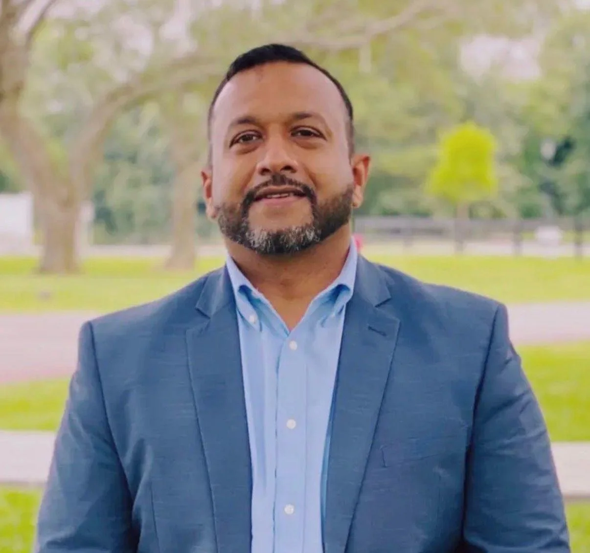 A man in a blue suit and light blue shirt standing outdoors in a park with trees and grass in the background.
