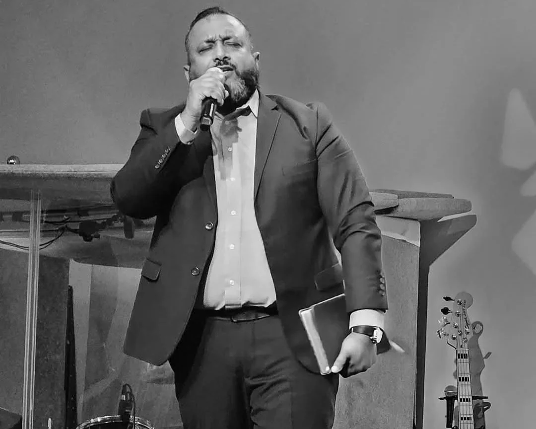 A man in a suit holding a microphone and a book or notebook, singing or speaking with his eyes closed.