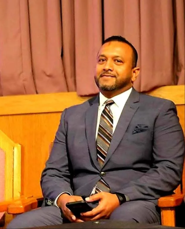 A man in a dark suit and striped tie sitting on a wooden chair, holding a smartphone, with pink curtains in the background.