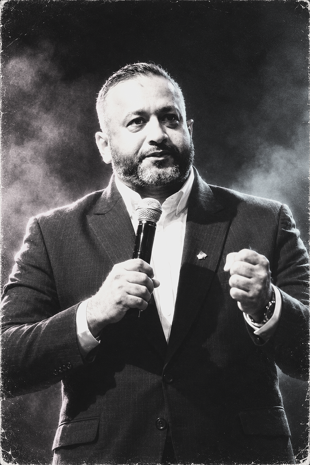 A man with a beard and mustache holding a microphone and speaking, wearing a suit and a small American flag pin on his lapel.