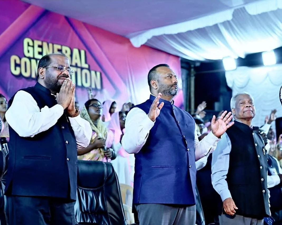 Three men standing at a church service with their right hands raised, attending a General Convocation. The background has people with their hands raised and a large pink and purple banner that reads 'GENERAL CONVOCATION.'