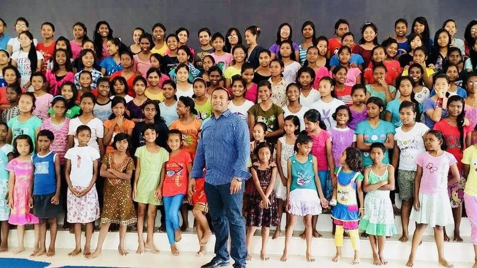 A large group of children and a man posing for a photo indoors, with a gray backdrop, many children are smiling and wearing colorful clothes.