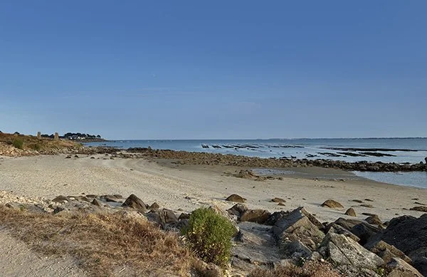 Plage de sable avec des rochers, vue sur la mer sous un ciel bleu clair.