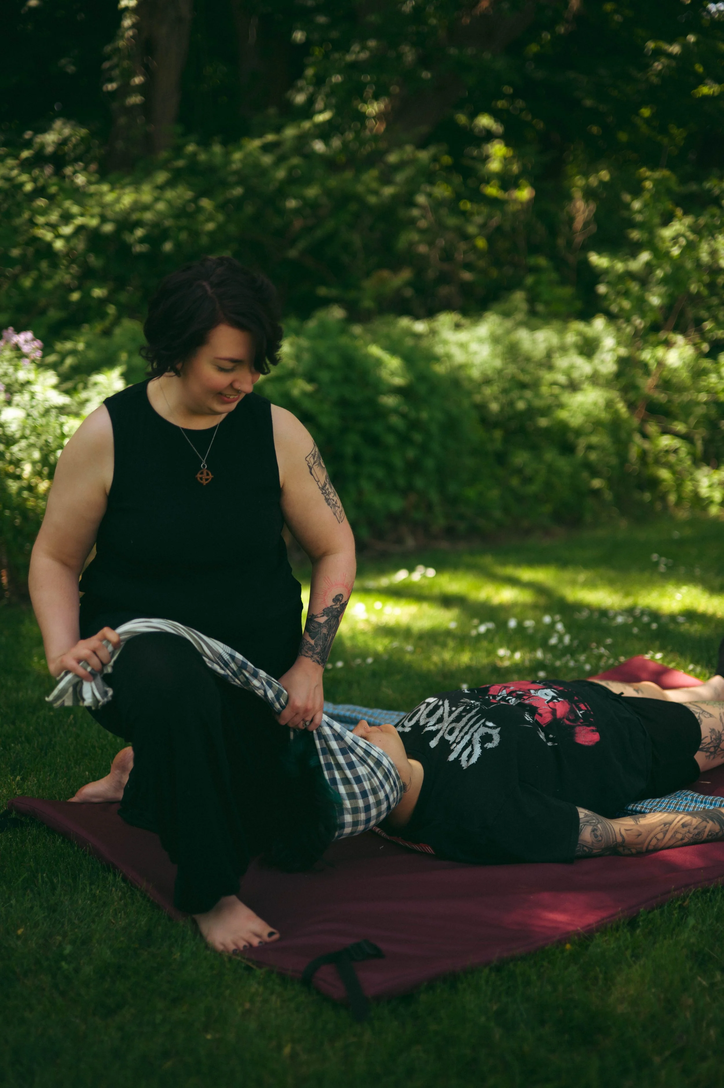 A woman with tattoos and dark curly hair practicing a yoga pose outdoors on a mat, with another person lying face down on the mat in a park surrounded by green trees and grass.