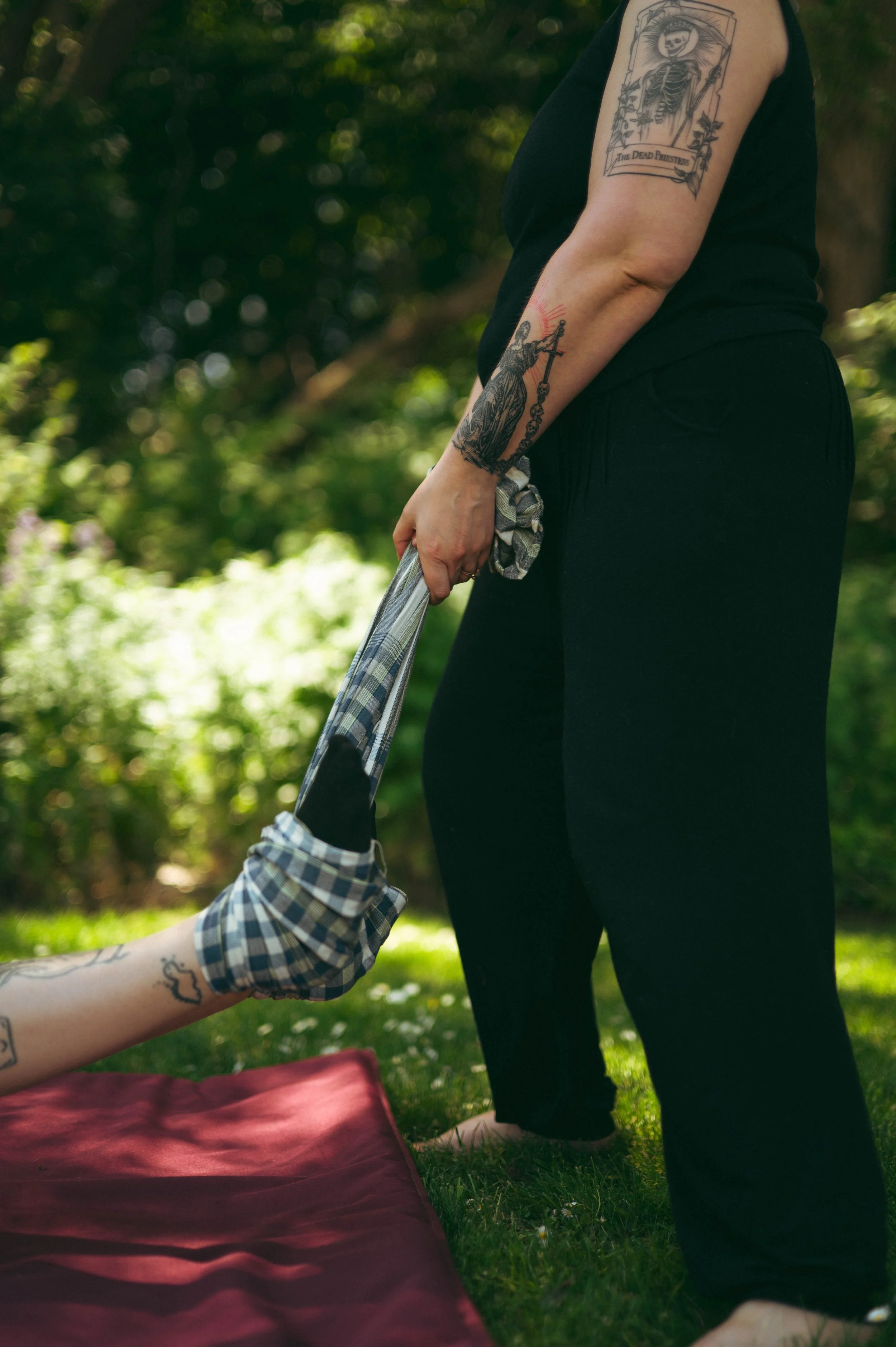 A person with tattoos on their arm standing outdoors in a park, holding the hand of a person sitting or lying on the grass, with a blanket. The person standing has tattoos including a skeleton in a coffin on their upper arm and a figure holding a sword on their forearm. The grassy area and trees are visible in the background.