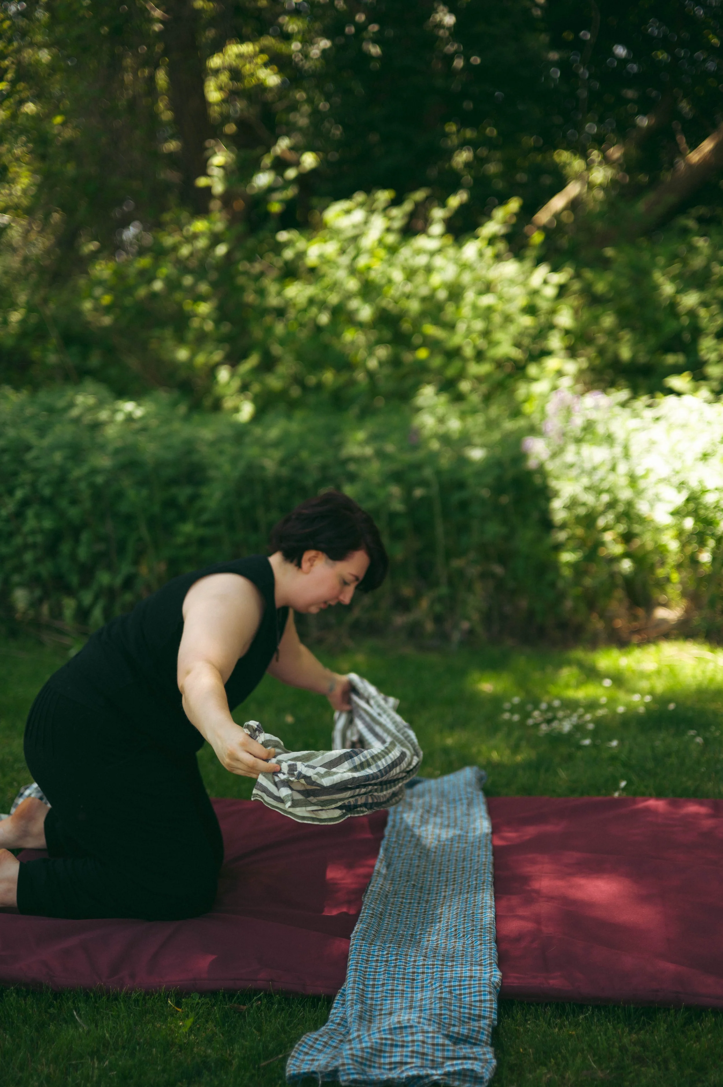 A woman is crouching down outdoors, folding a plaid blanket into a bag, with a blanket spread on the grass in front of her. There are trees and greenery in the background under bright sunlight.
