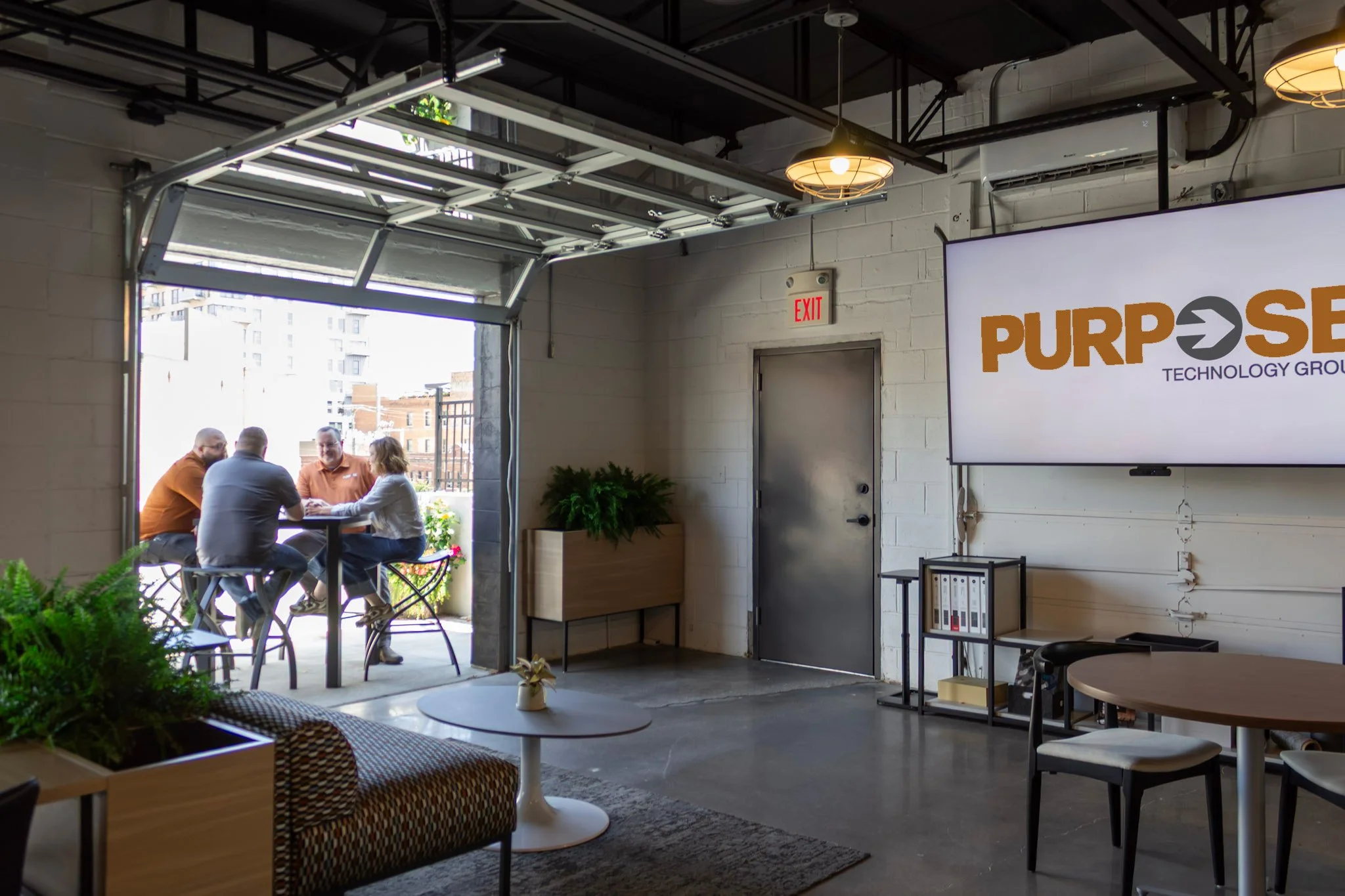 Indoor meeting space with a group of four people sitting at a table outside, viewed through an open garage-style door. The room has modern decor, plants, and a large screen displaying the logo of 'PURPOSE TECHNOLOGY GROUP' on a white wall.