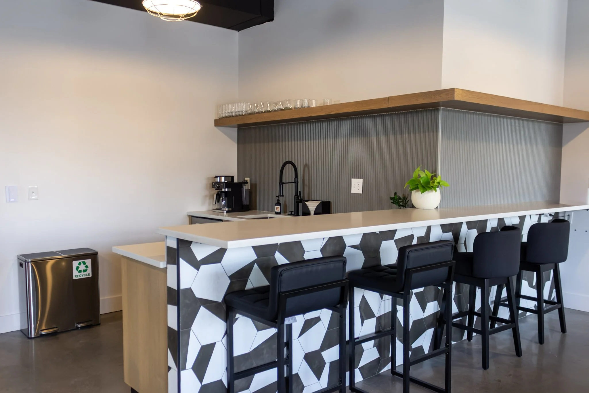 A modern kitchen with a black, white, and grey hexagon patterned island, black chairs, a wooden shelf with glasses, a coffee machine, black faucet, potted plant, and a recycle bin.