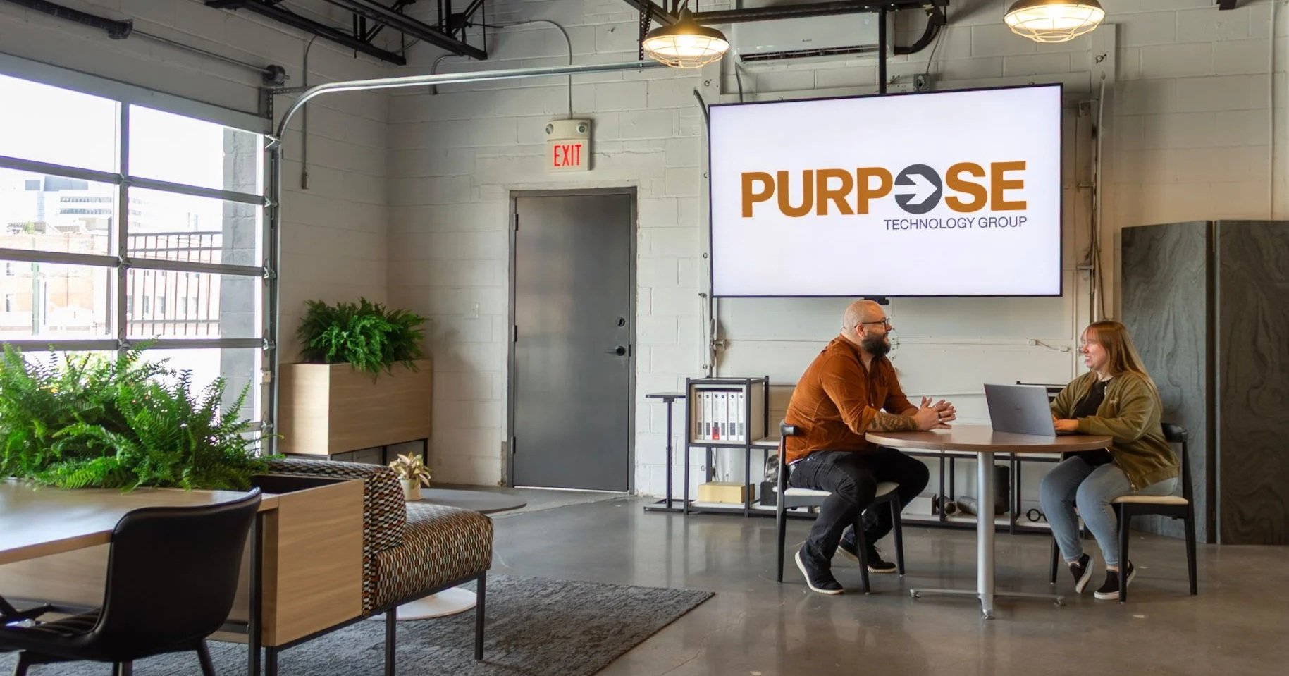 Two people meeting in a modern office event space with garage doors. A large screen on the wall reads Purpose Technology Group.
