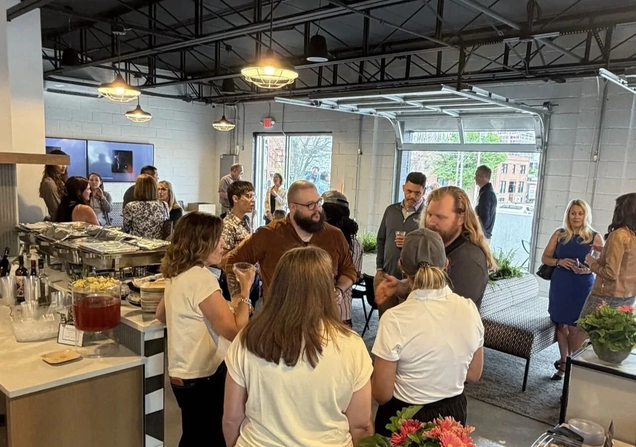 People socializing at a gathering or party in an industrial-style room with exposed ceilings and large garage doors, some near a bar area with bottles and snacks.