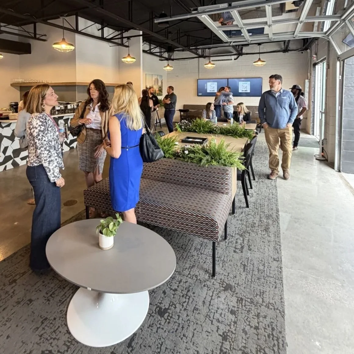 Indoor gathering with people talking and networking in a modern, open-concept space with a high ceiling, black and white decor, and digital screens on the wall.