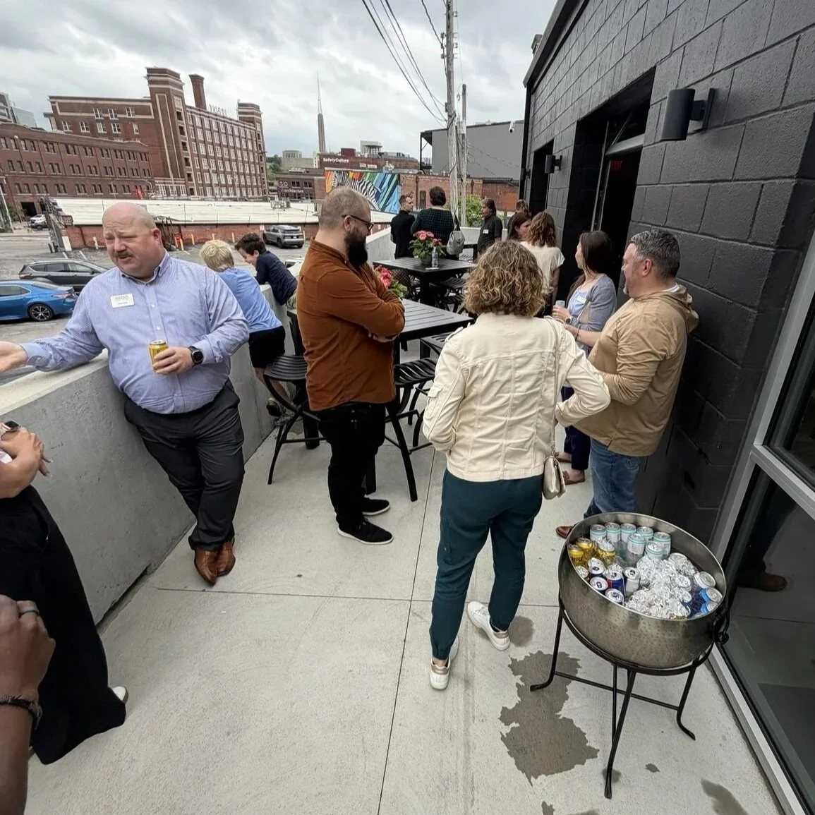 People gathered on a rooftop terrace with a black brick wall, drinking beverages, and socializing under a cloudy sky. A table with cans of drinks and foil-wrapped items is visible. The city skyline with buildings and power lines can be seen in the ba