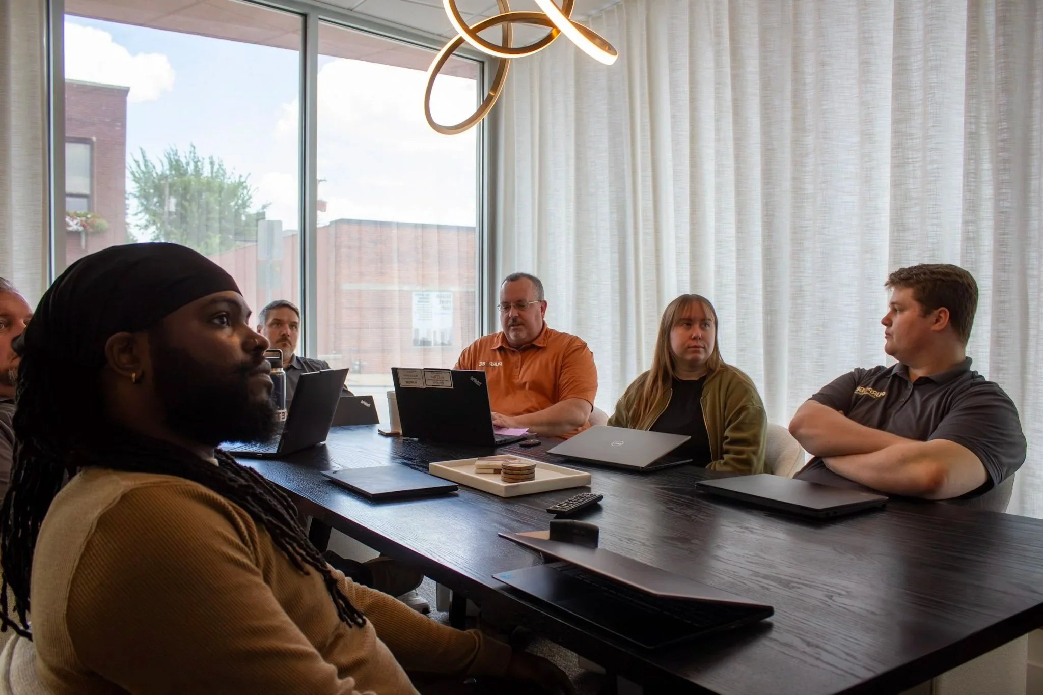 A diverse group of people sitting around a conference table in a modern meeting room with large windows and white curtains, engaged in discussion or listening attentively.