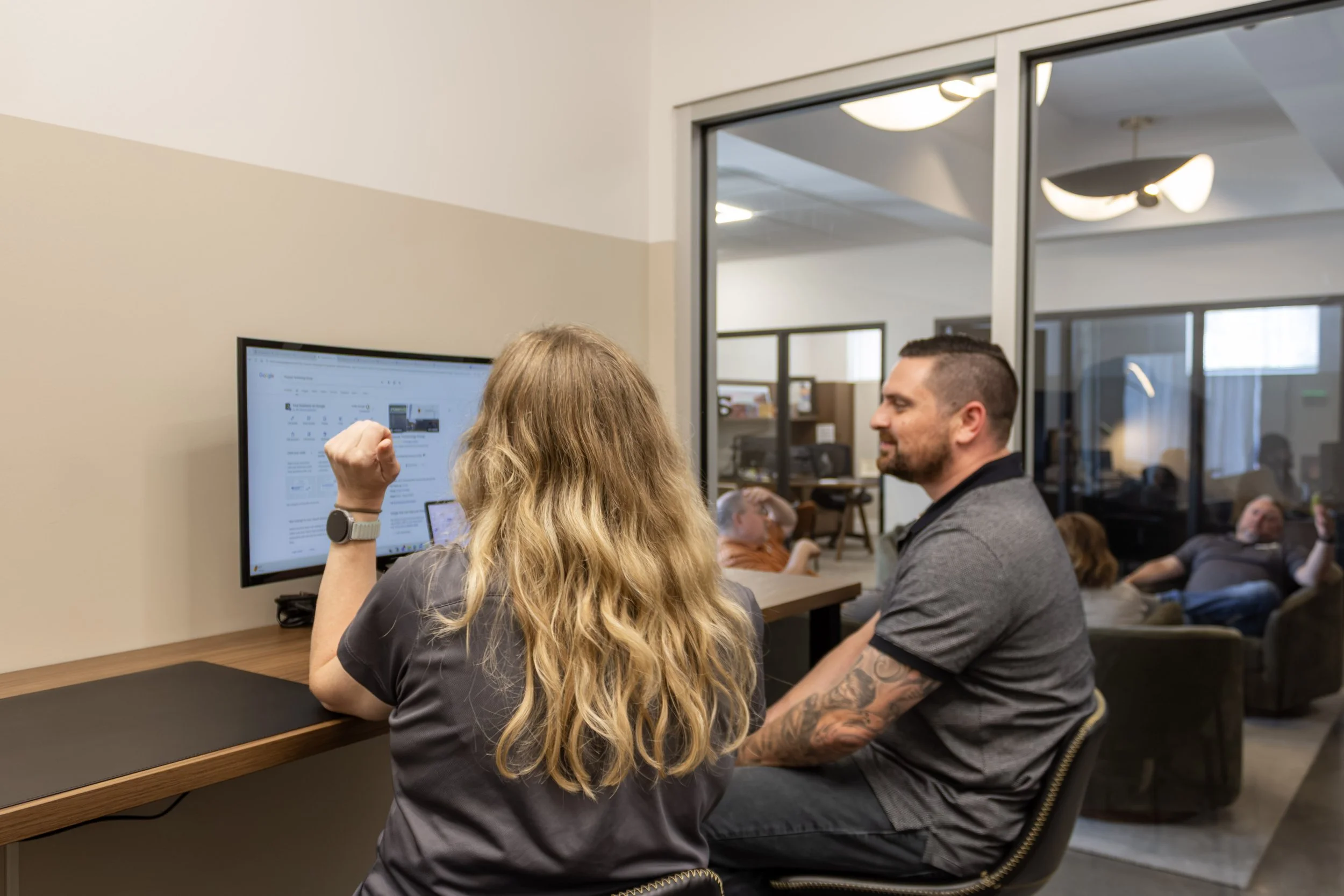 A woman with long blonde hair wearing a smartwatch on her wrist, fist raised, sitting at a desk in front of a computer monitor. A man with short hair and tattoos on his arm, wearing a gray shirt, is sitting across from her, smiling and engaged in conversation. In the background, there are people sitting and a ceiling with modern lighting fixtures.