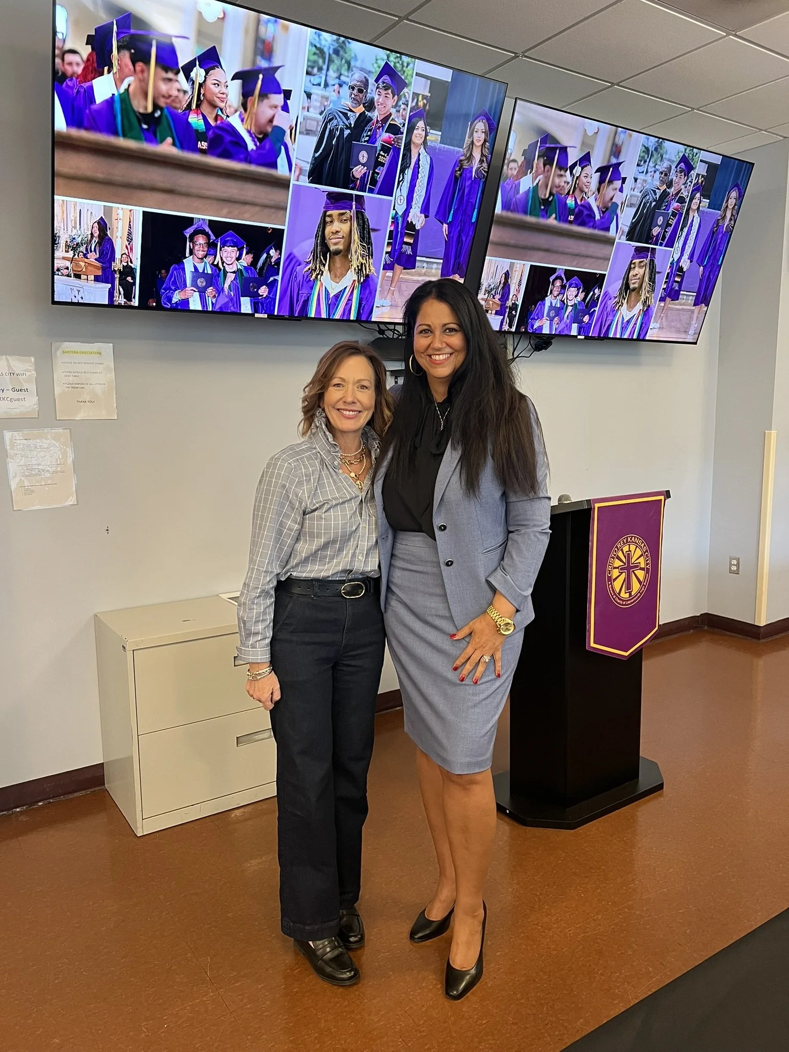 Two women standing together at a graduation event, smiling at the camera, with a display of graduation photos in the background.