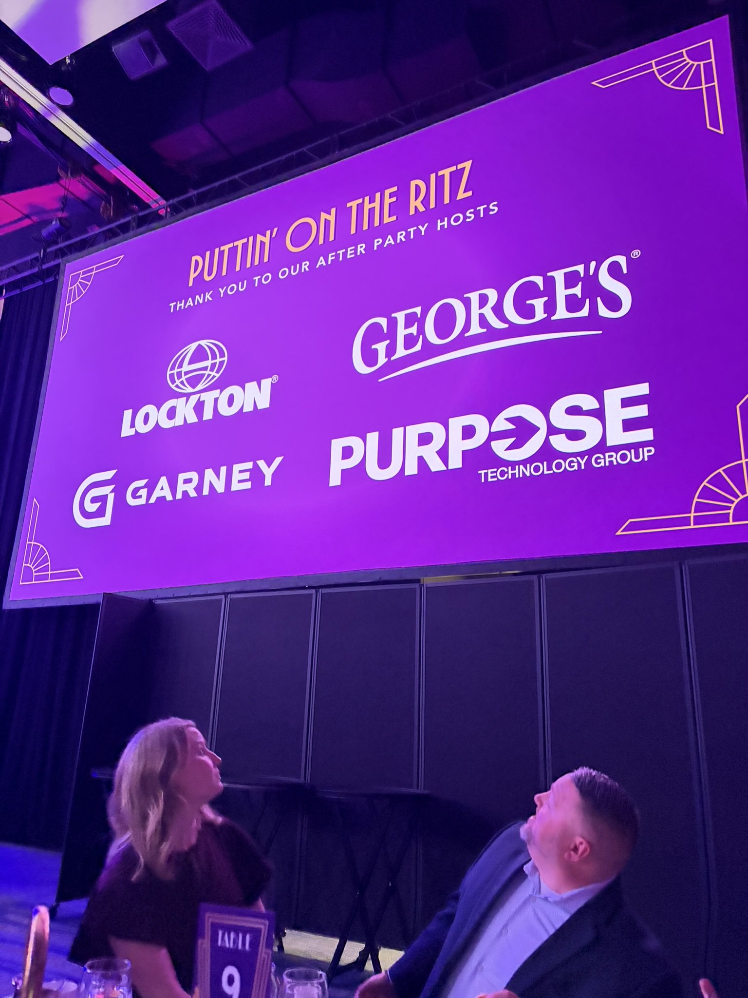 Event with a large purple and yellow screen displaying the text 'Puttin' On The Ritz' and logos for Lockton, Georges, Garney, and Purpose. Attendees are seated at tables in front of the screen.