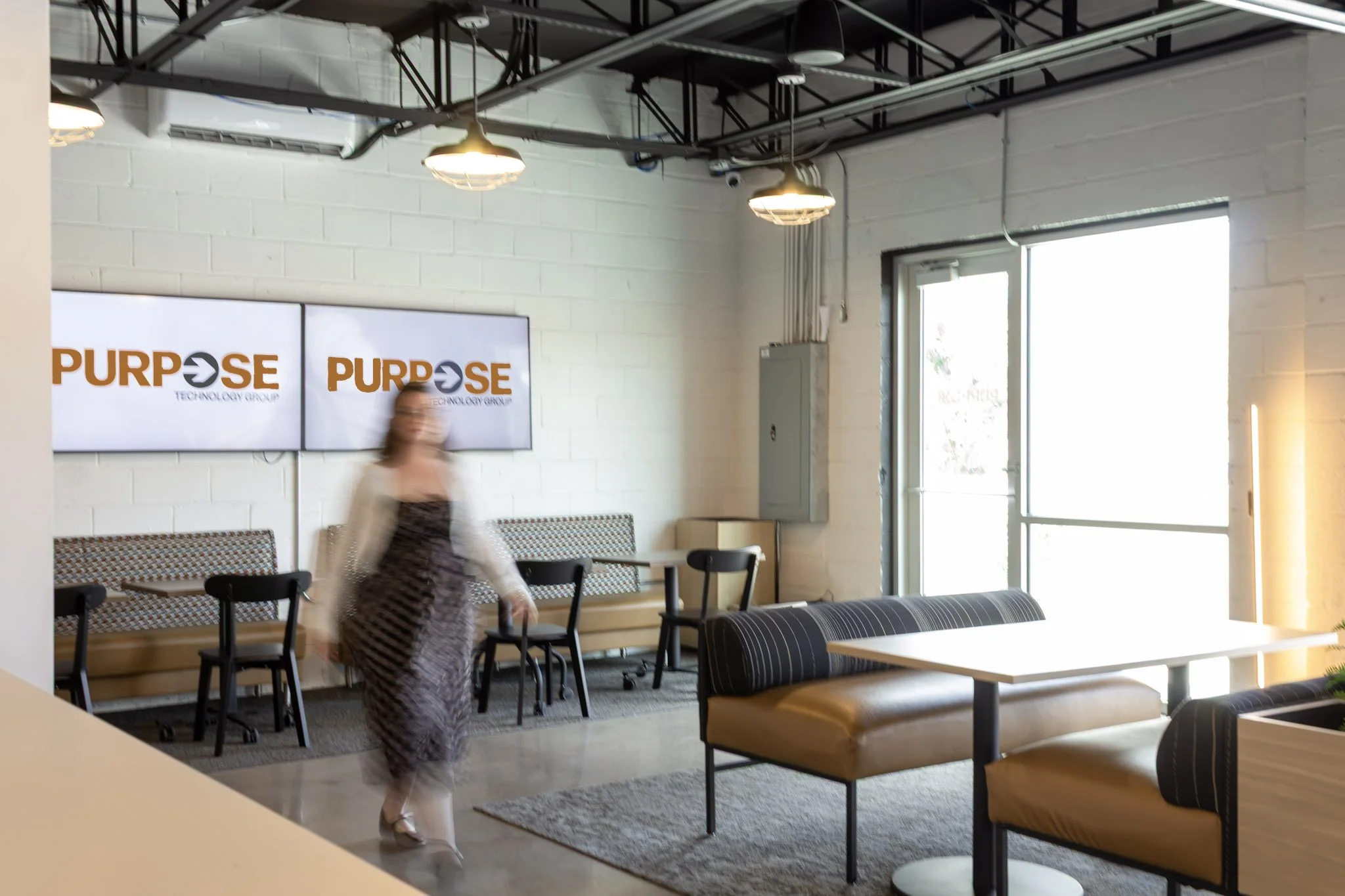 A picture of a women walking, she is blurred in a modern office with Purpose Technology Group logos on two wall screens behind her.