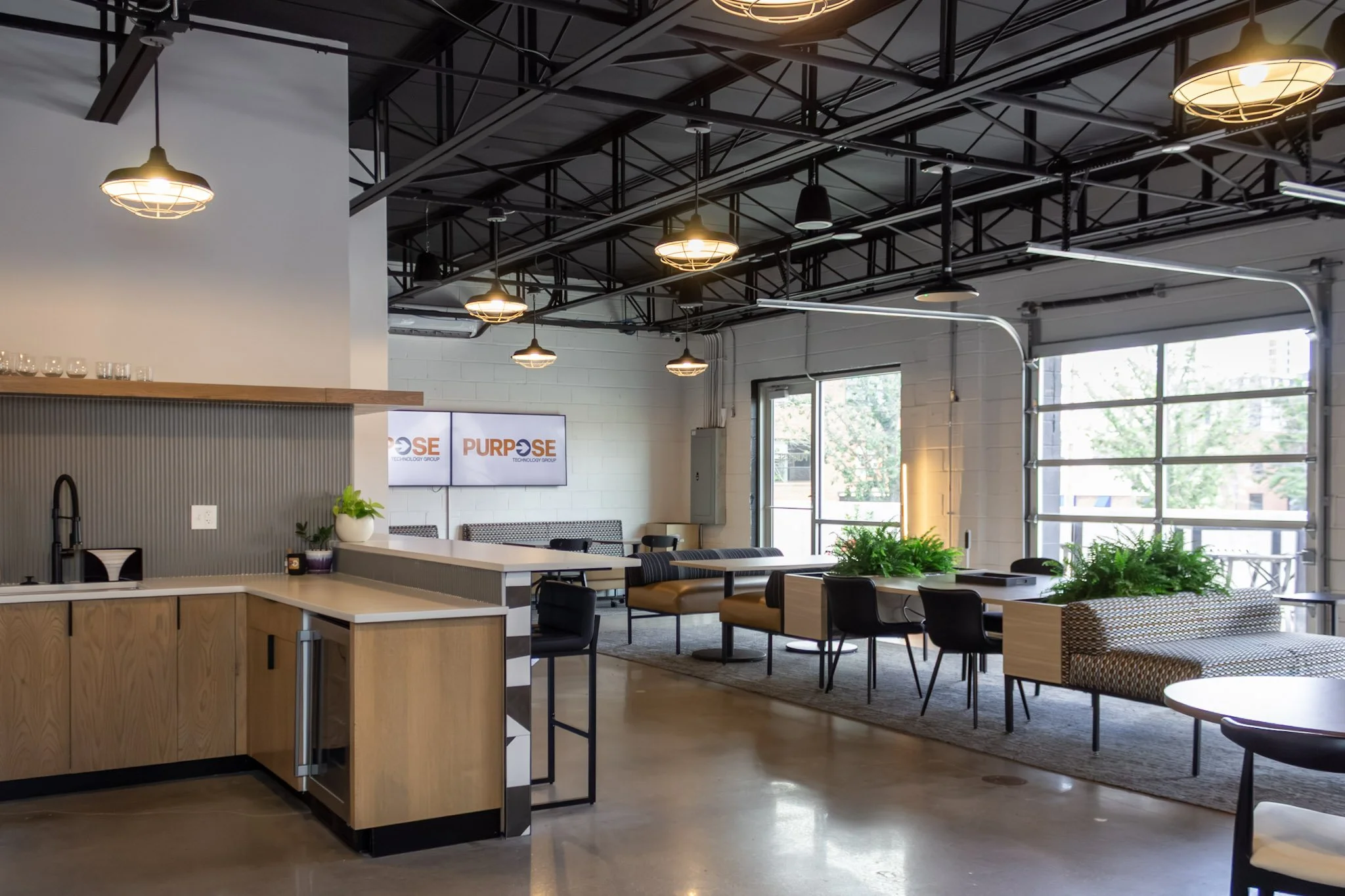 Interior of a modern office or co-working space with industrial ceiling lights, large windows, and a mix of dining tables and lounge seating, with some greenery and a wall-mounted TV displaying the logo 'PURPOSE'.