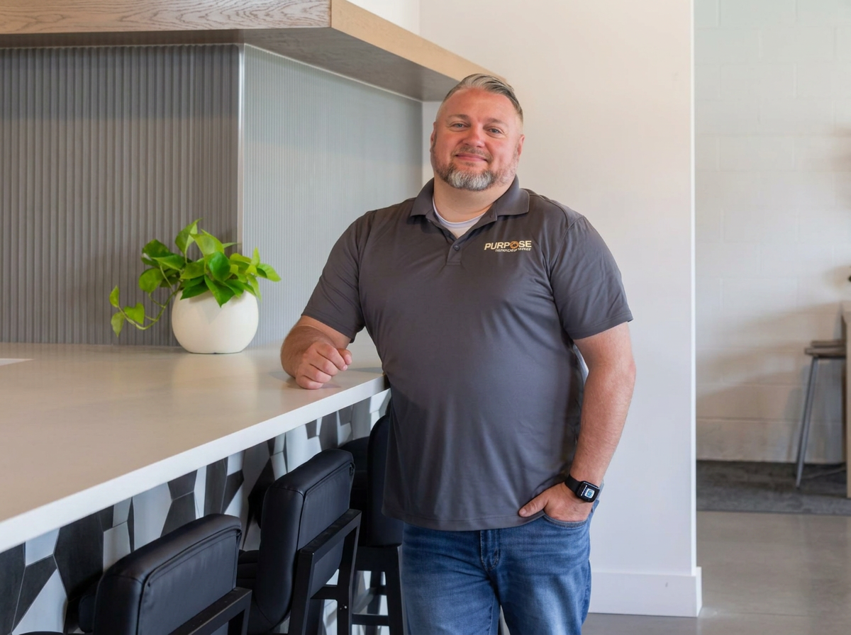 A man with light hair and beard wearing a gray polo shirt and jeans, standing in a modern space, leaning on a white counter with a potted green plant.