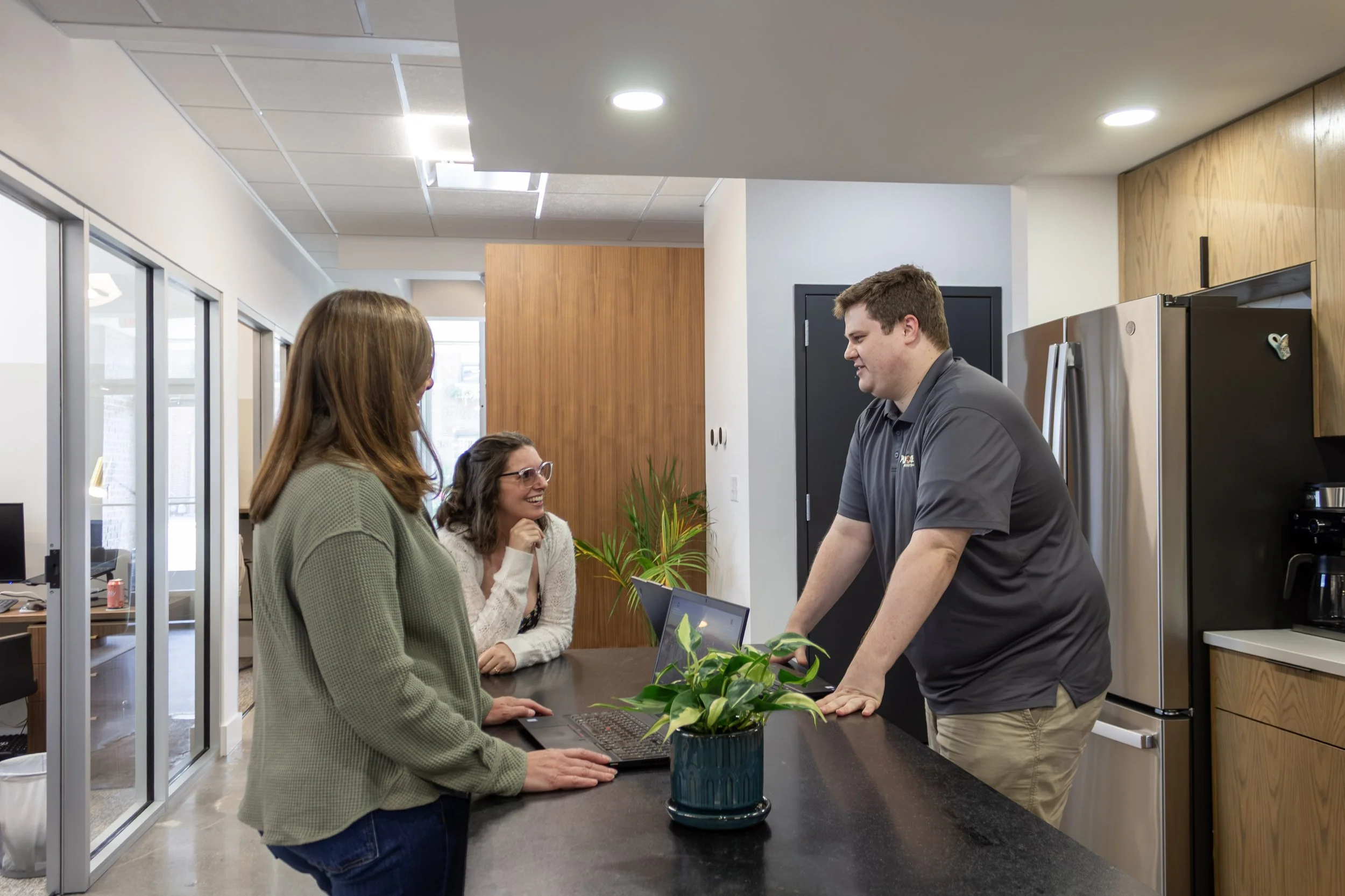 Four people in an office or reception area, two women and two men, engaged in conversation around a counter with a laptop and potted plant, with modern decor and large windows in the background.
